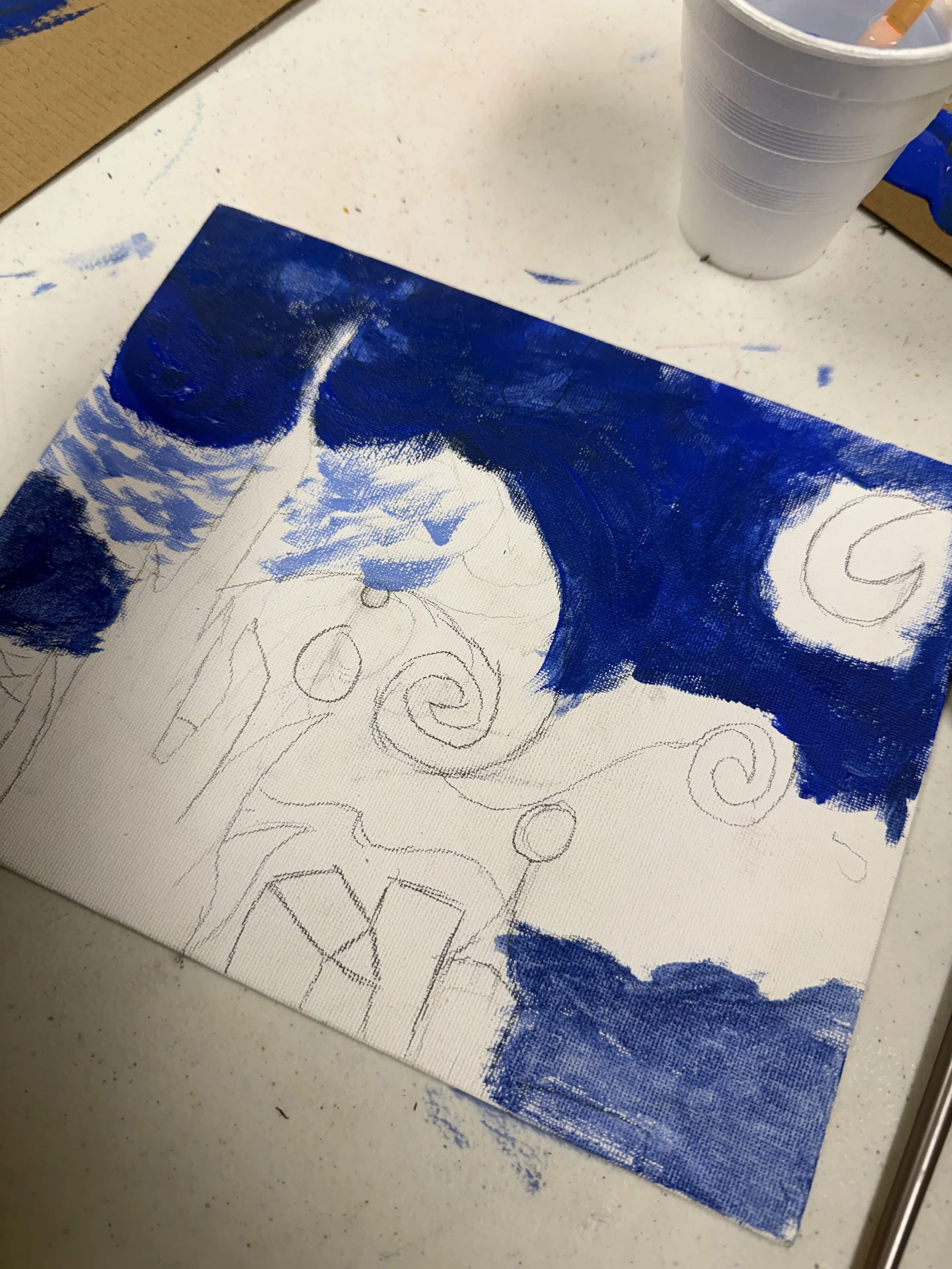 A canvas with a partially painted blue and white abstract design, with pencil sketches underneath, on a white table next to a stack of plastic cups.