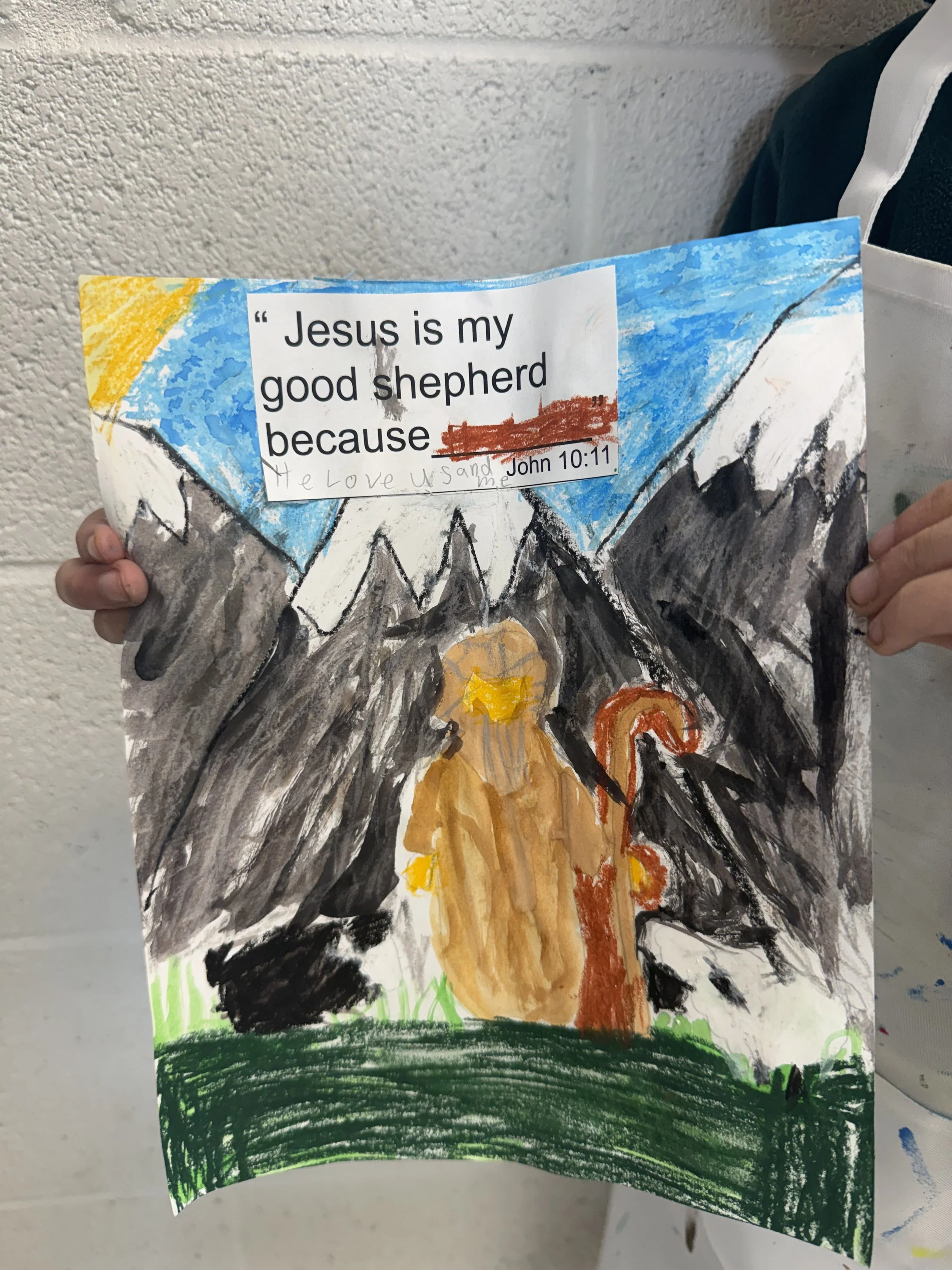 Child's colorful drawing depicting a mountain landscape with snow-capped peaks, a sunny sky, a figure with a halo and a staff, and a patch of green grass. A religious quote sticker reads, "Jesus is my good shepherd because" with John 10:11 citation.