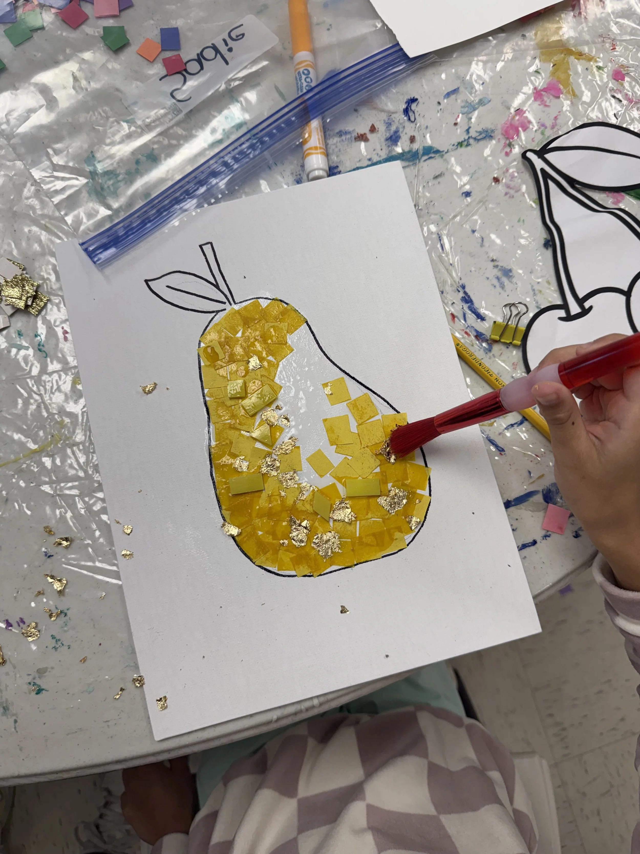 A child is creating a mosaic art project of a pear on a white canvas. The pear outline is drawn in black marker, and the child is gluing yellow and gold foil pieces inside the outline. The workspace is covered with various art supplies and gold foil 