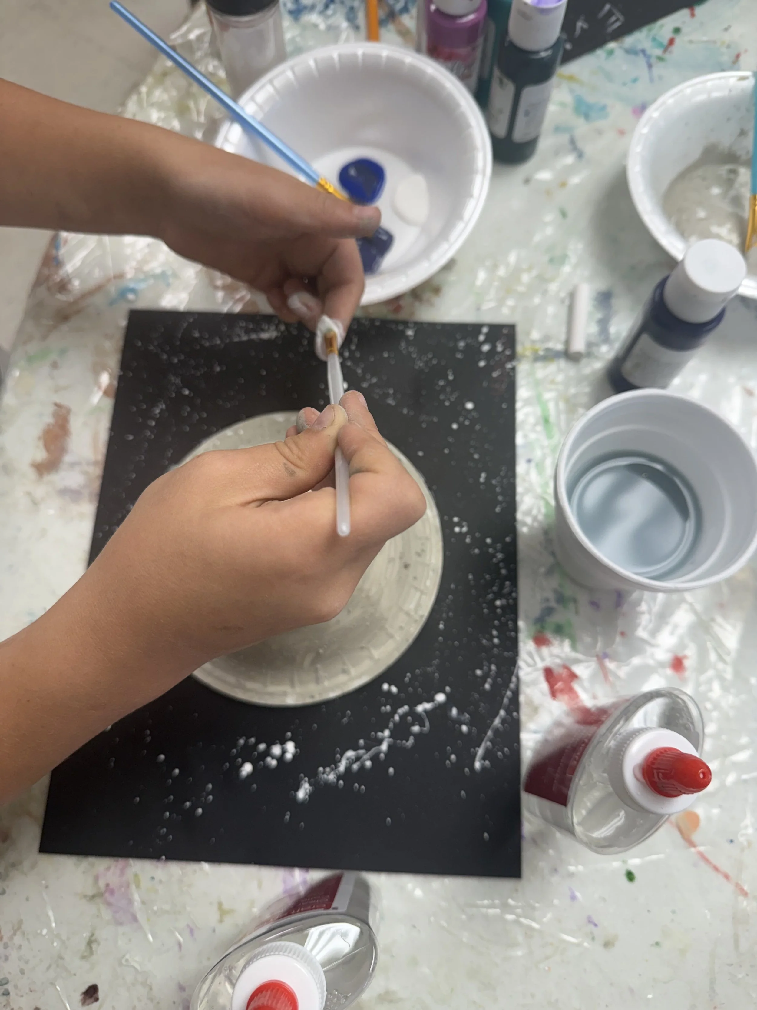 Close-up of hands pouring paint onto a black canvas, with various bottles of paint and art supplies on a table covered in paint splatters.