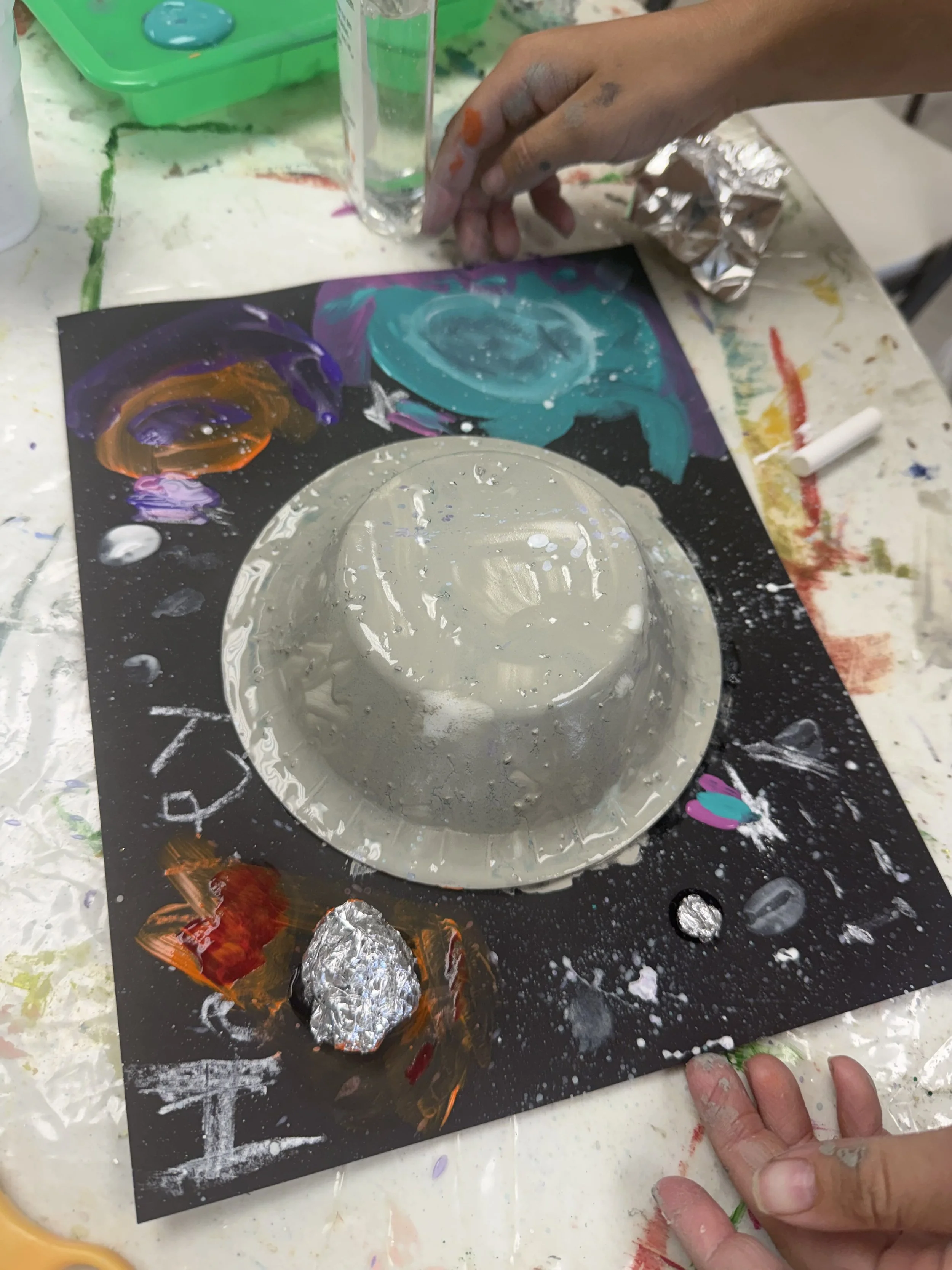 A person creating a solar system model with painted foam cups on a black paper background decorated with colorful galaxy designs and planets, surrounded by art supplies and paint stains.