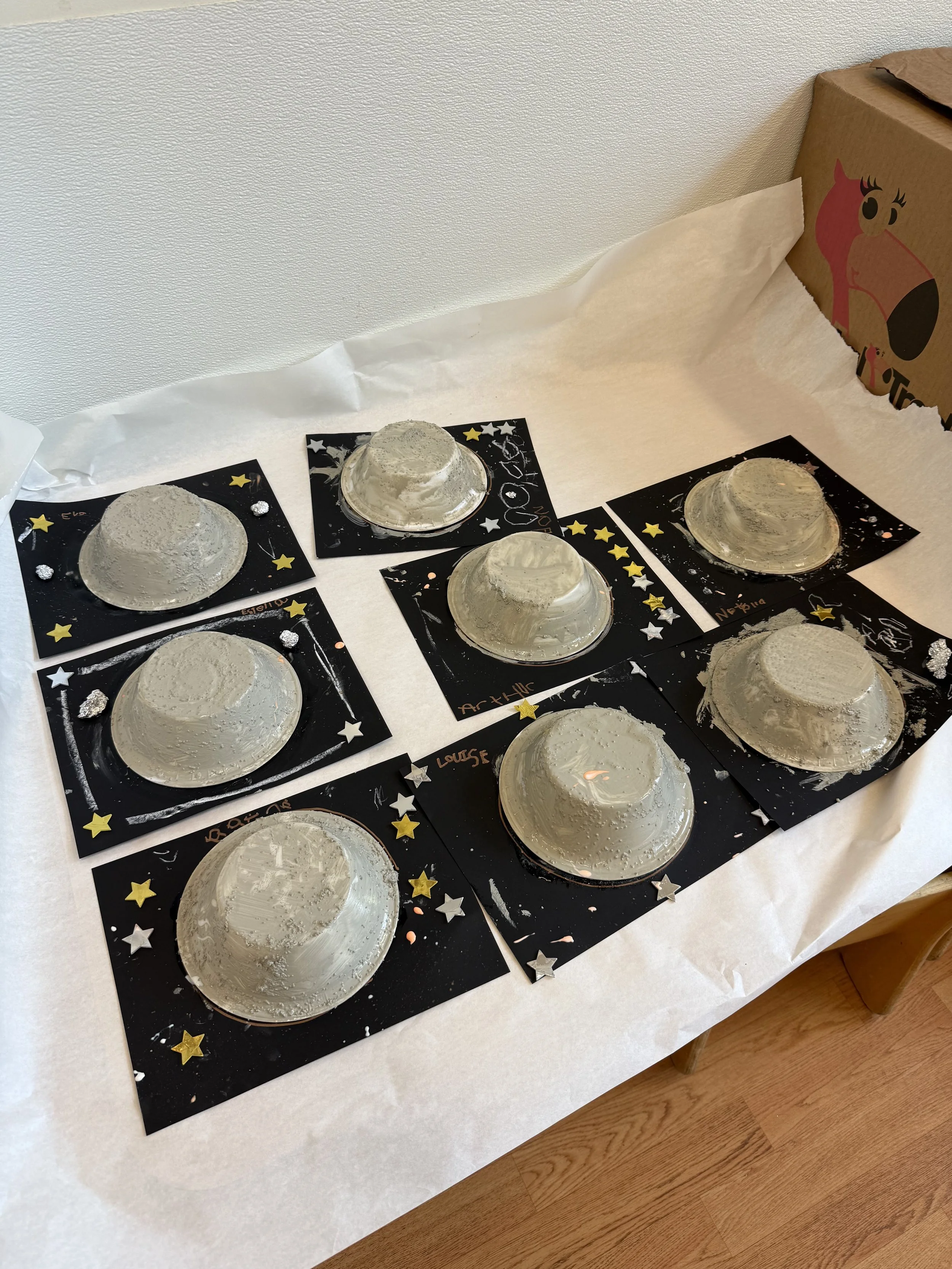 Six paper mache bowls painted silver, each on a black decorated paper with stars and personalized names, arranged on a white table with tissue paper, and a cardboard box with a horse illustration in the background.