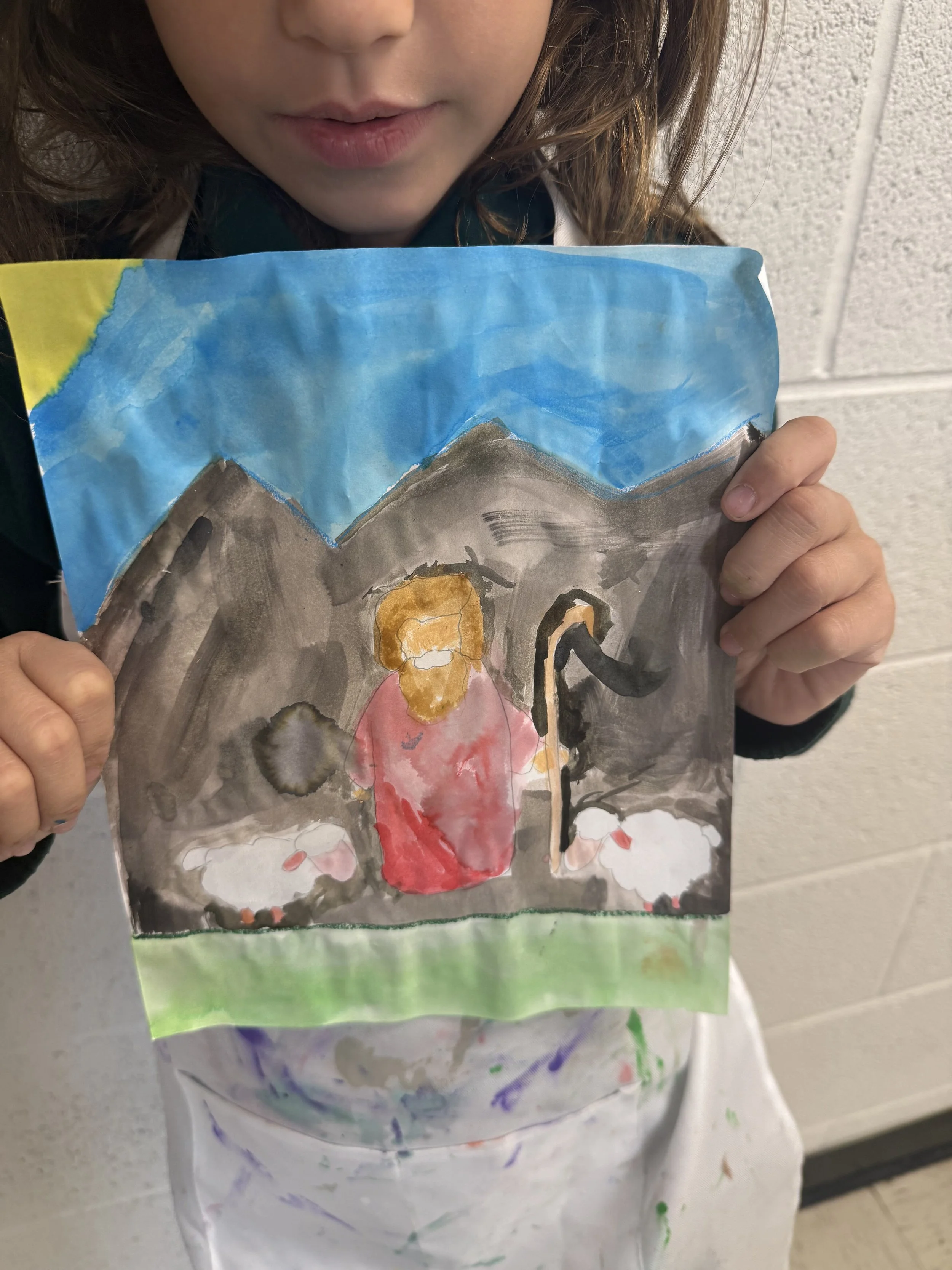 Child holding a colorful drawing of a nativity scene with a figure in red, sheep, and mountains in the background.