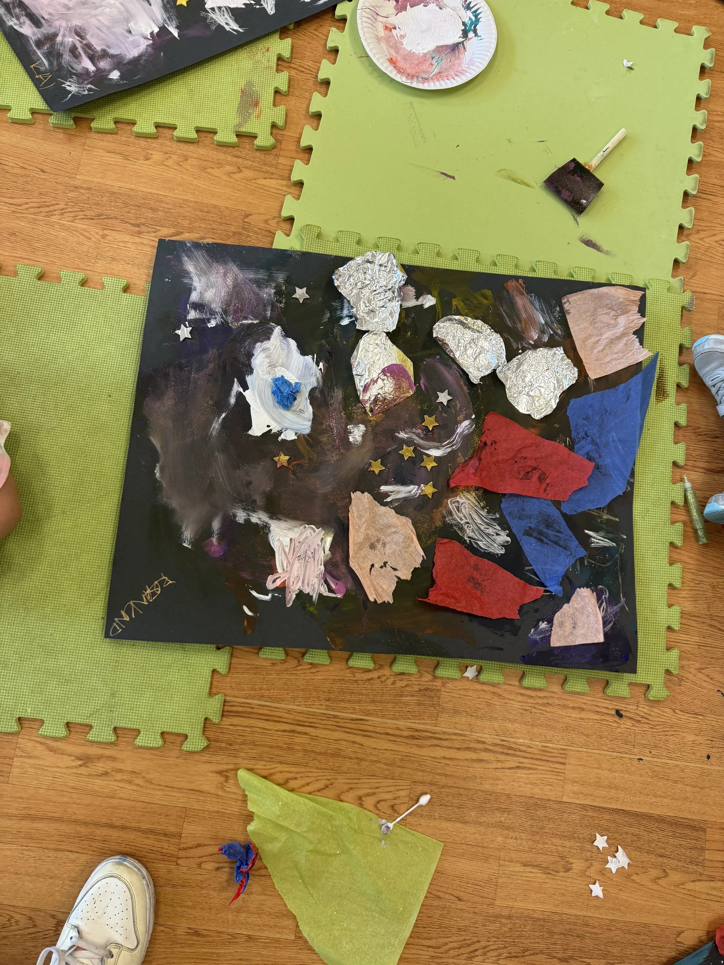 Child's art project on black paper with painted patches and torn colored paper, surrounded by green mats, a small plate with white paint, and scattered star-shaped confetti on a wooden floor.
