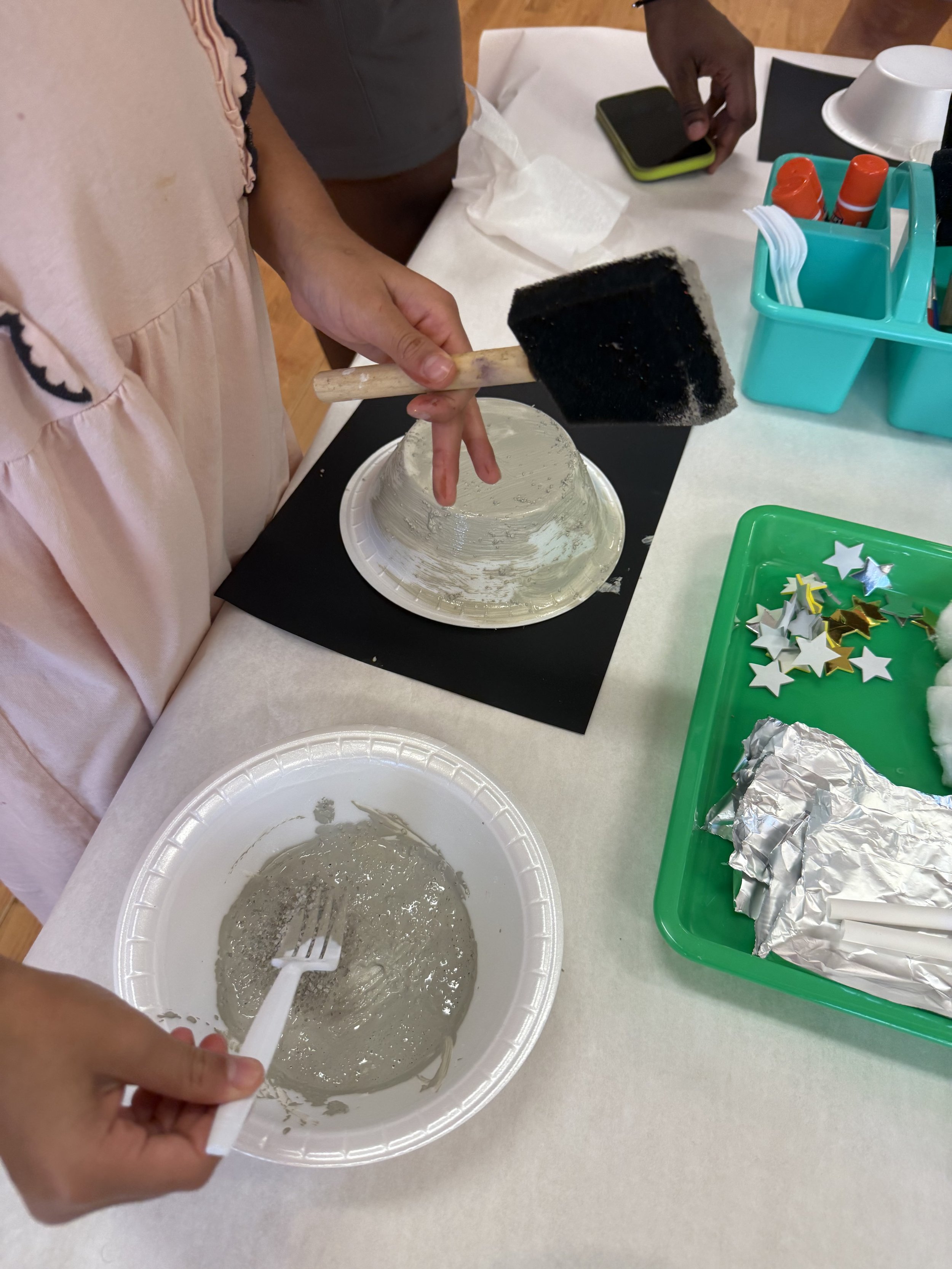 Children decorating a paper plate with glue, star-shaped confetti, and foil for a craft project.