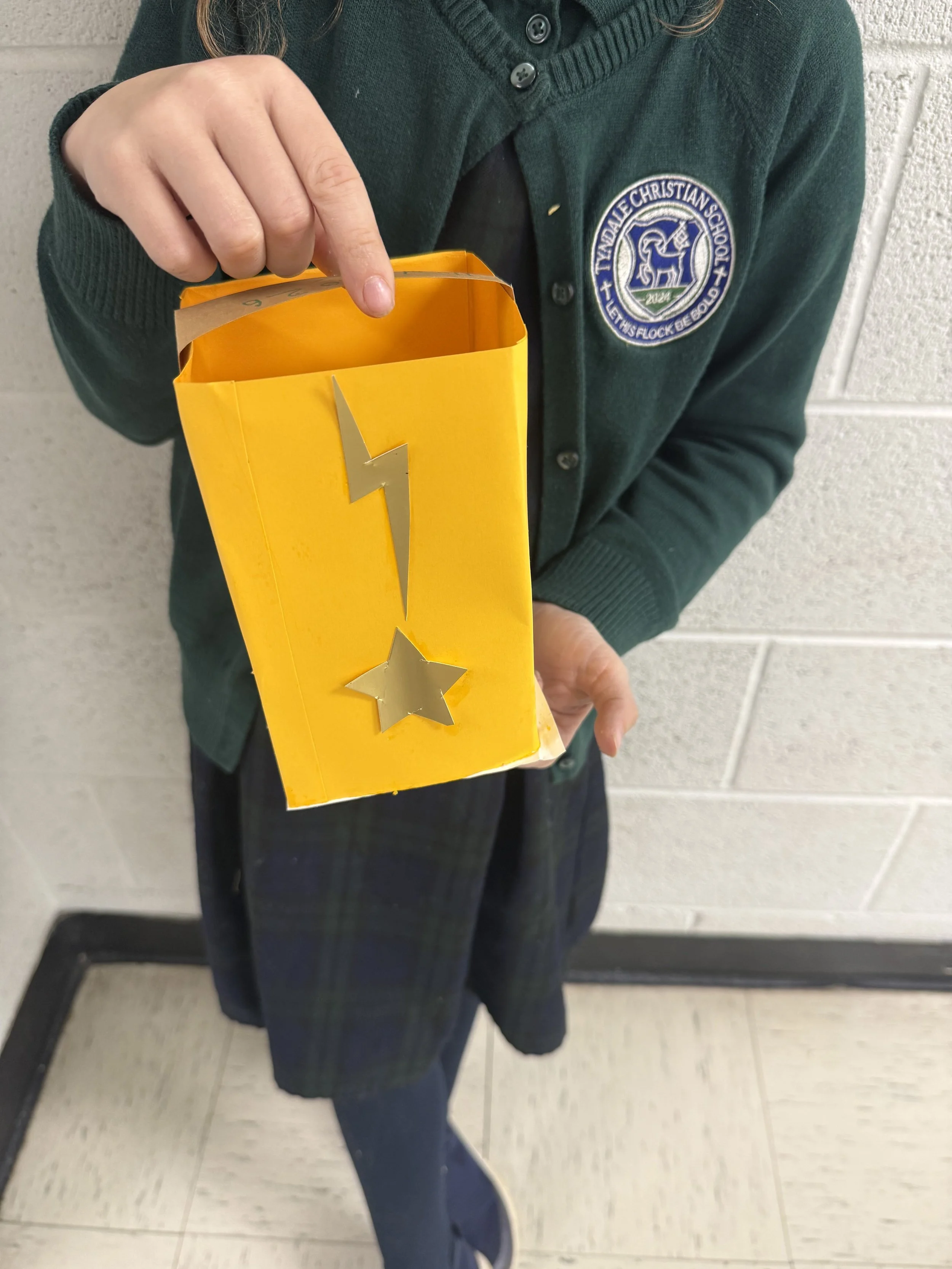 A student in a school uniform holding a yellow box with silver star and lightning bolt stickers on it, standing against a white brick wall.