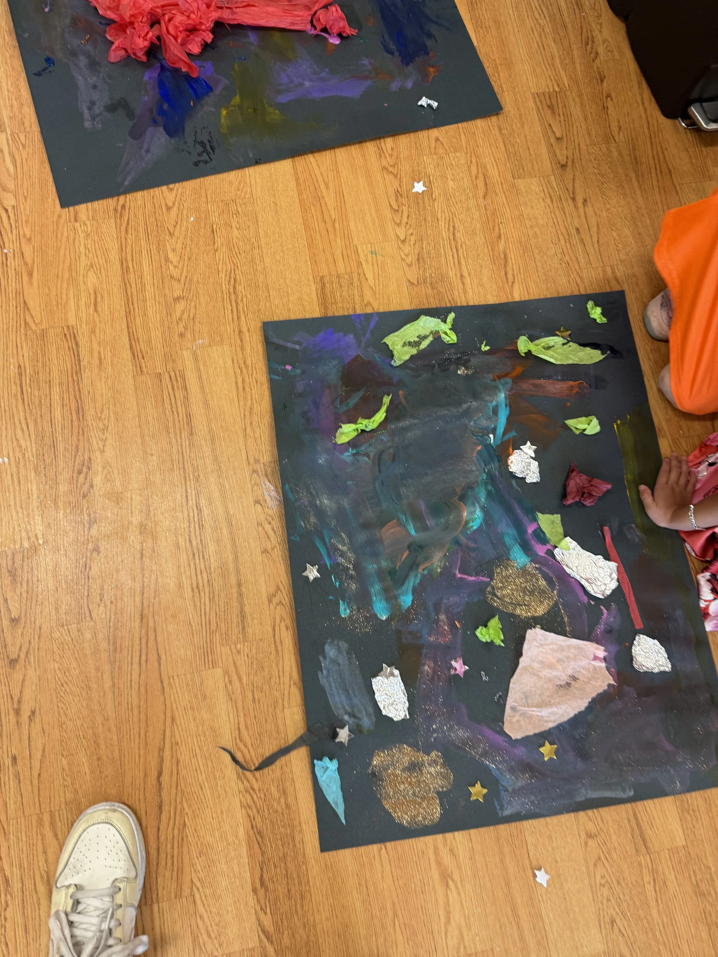 Colorful abstract children's art on black paper with torn tissue paper and star-shaped decorations on wooden floor.