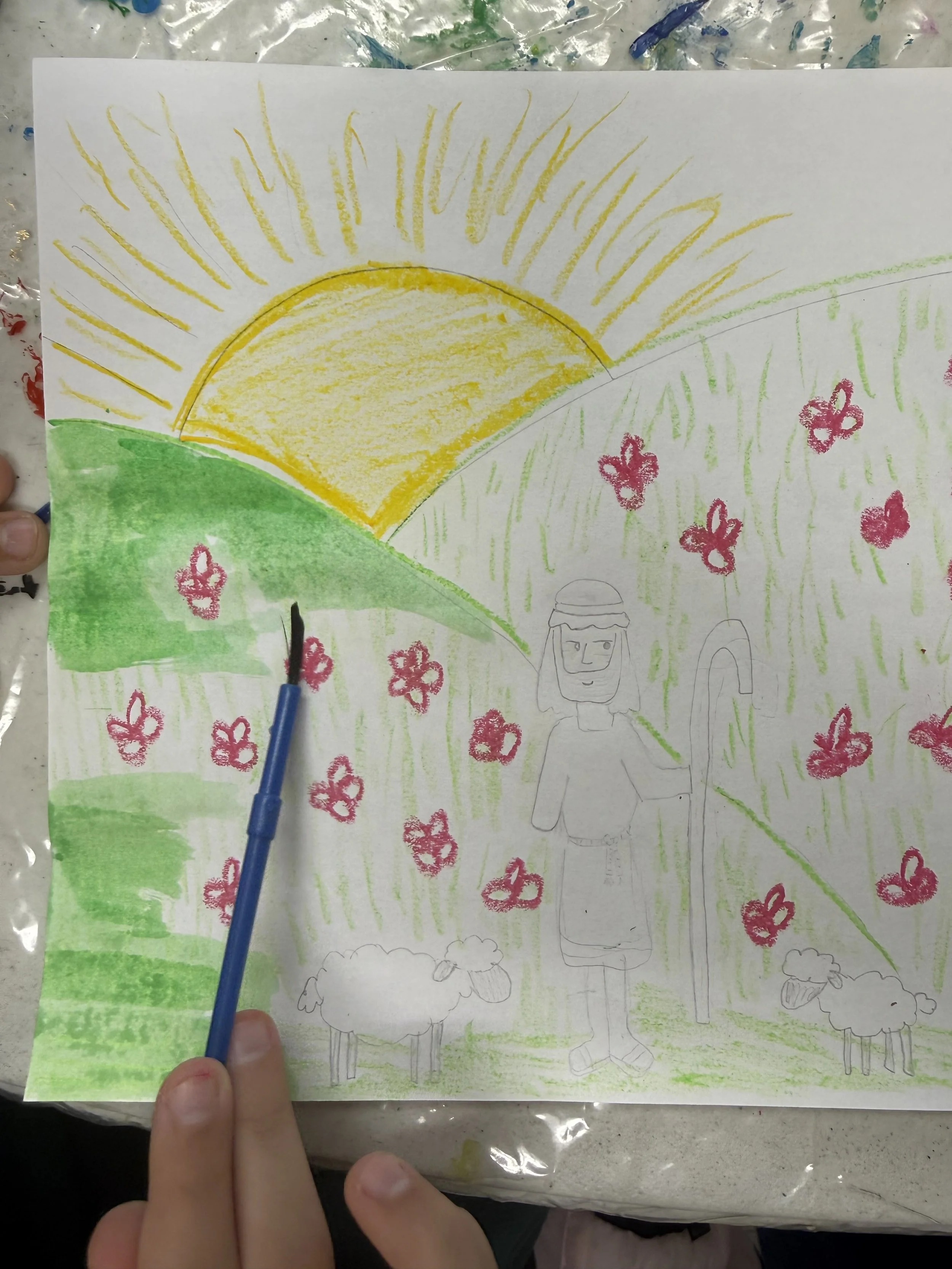 A children's drawing of a person standing in a green grassy field with pink flowers and sheep, with a bright yellow sun in the sky and green hilly terrain.