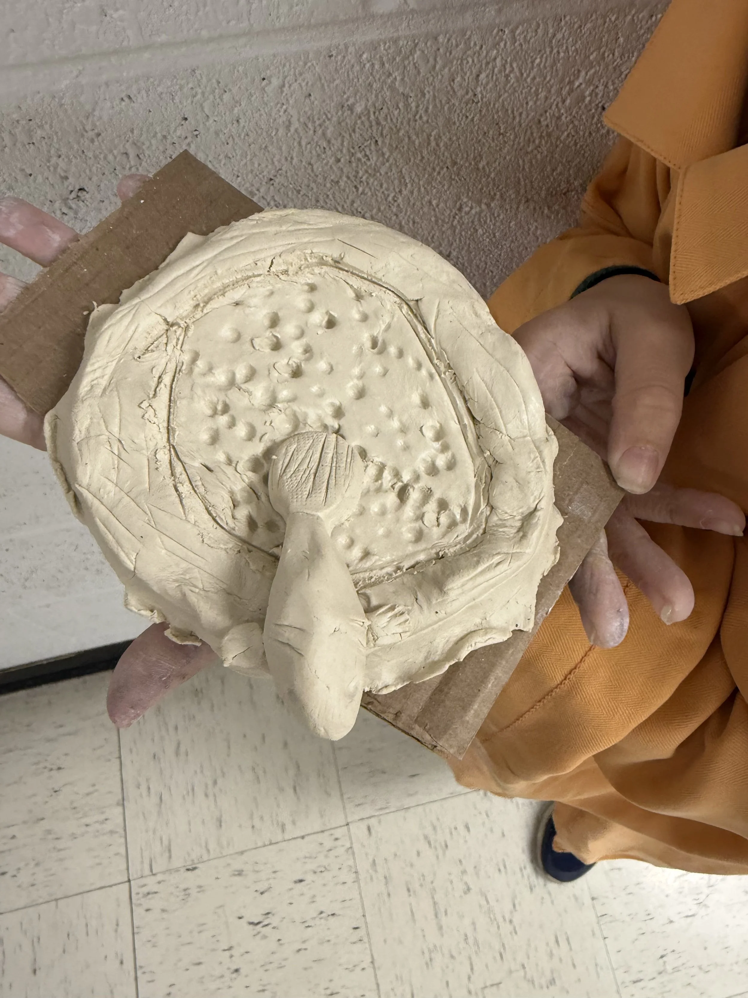 Unfinished pottery piece held by a person. The pottery appears to be a round, flat dish with a textured surface and a small, detailed raised figure in the center. The person is coated in clay on their hands.
