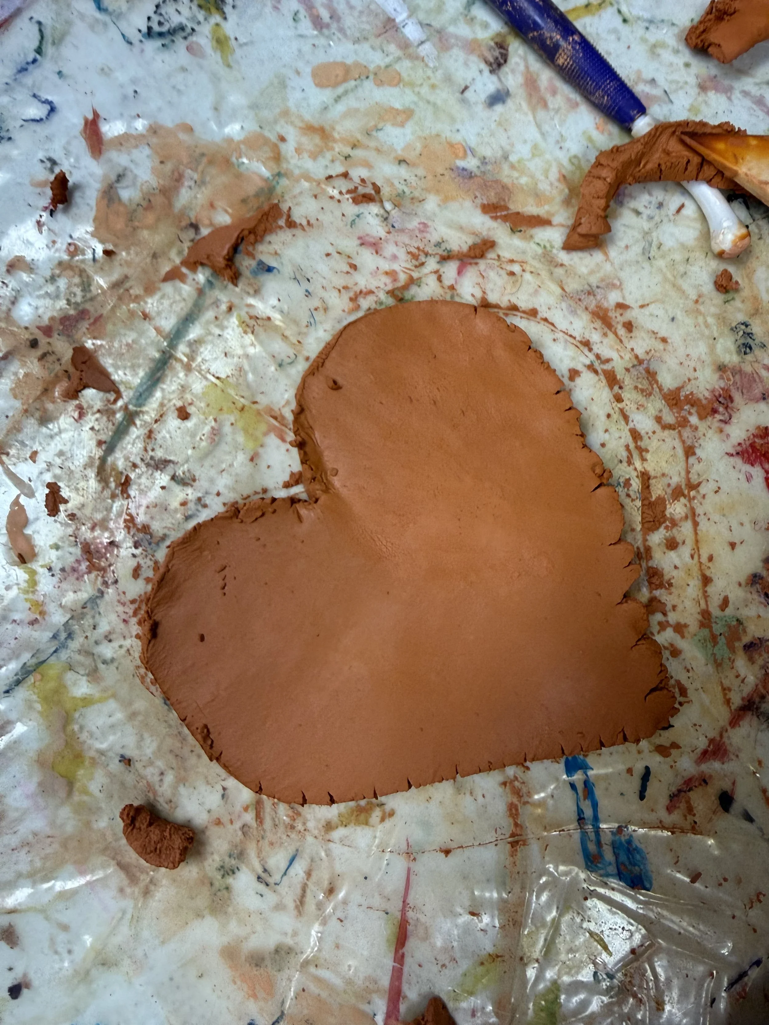 A pinkish-red clay heart shape placed on a colorful, messy surface with paint and clay scraps around it.