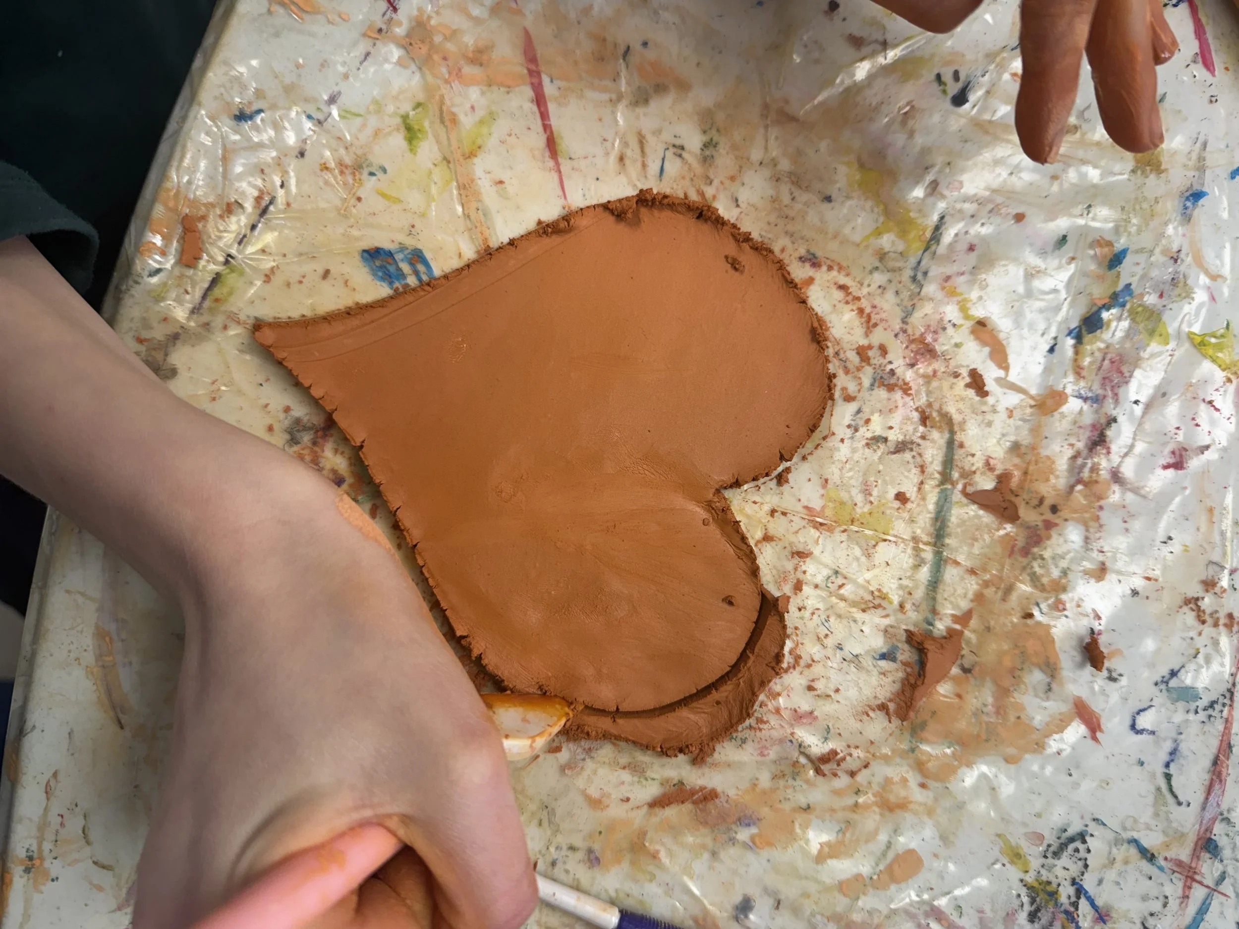 People shaping a large piece of brown clay on a messy, paint-splattered surface.