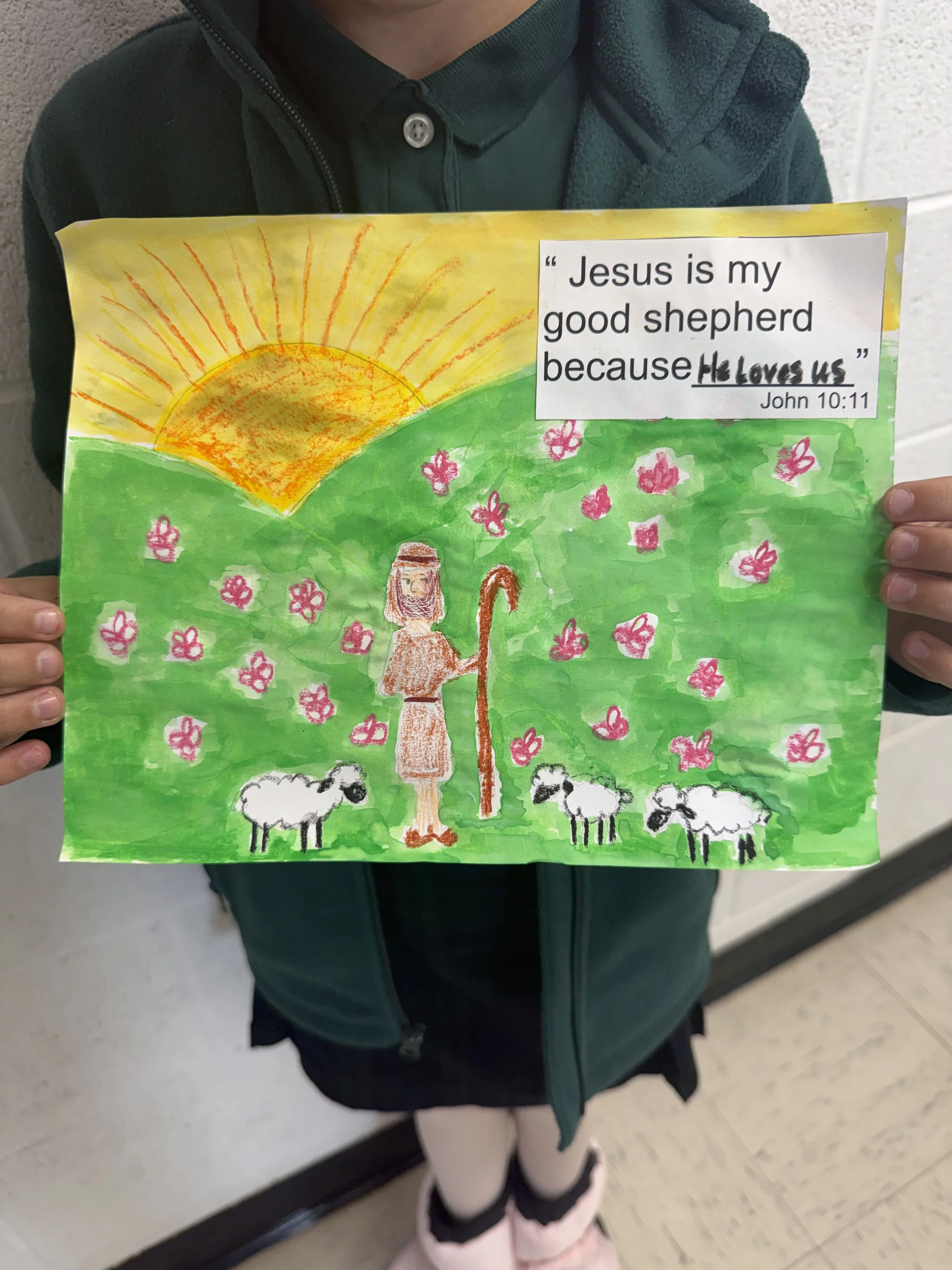 A child holds a colorful drawing of a shepherd in a green field with pink flowers, under a yellow sun with orange rays. There is a quote from John 10:11 about Jesus being a good shepherd.