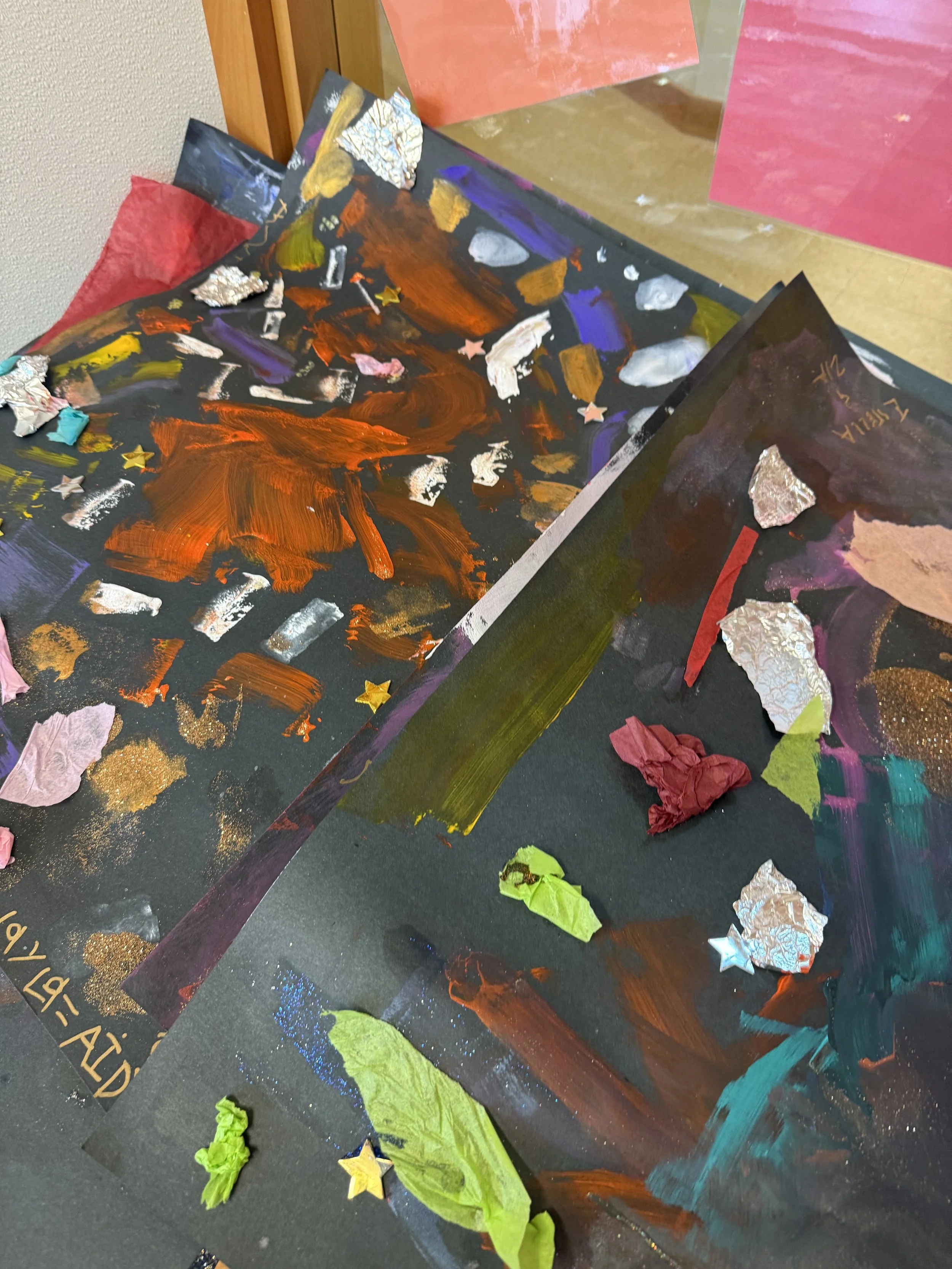 Colorful abstract paintings on black paper with torn tissue paper and star-shaped embellishments, displayed on a table.
