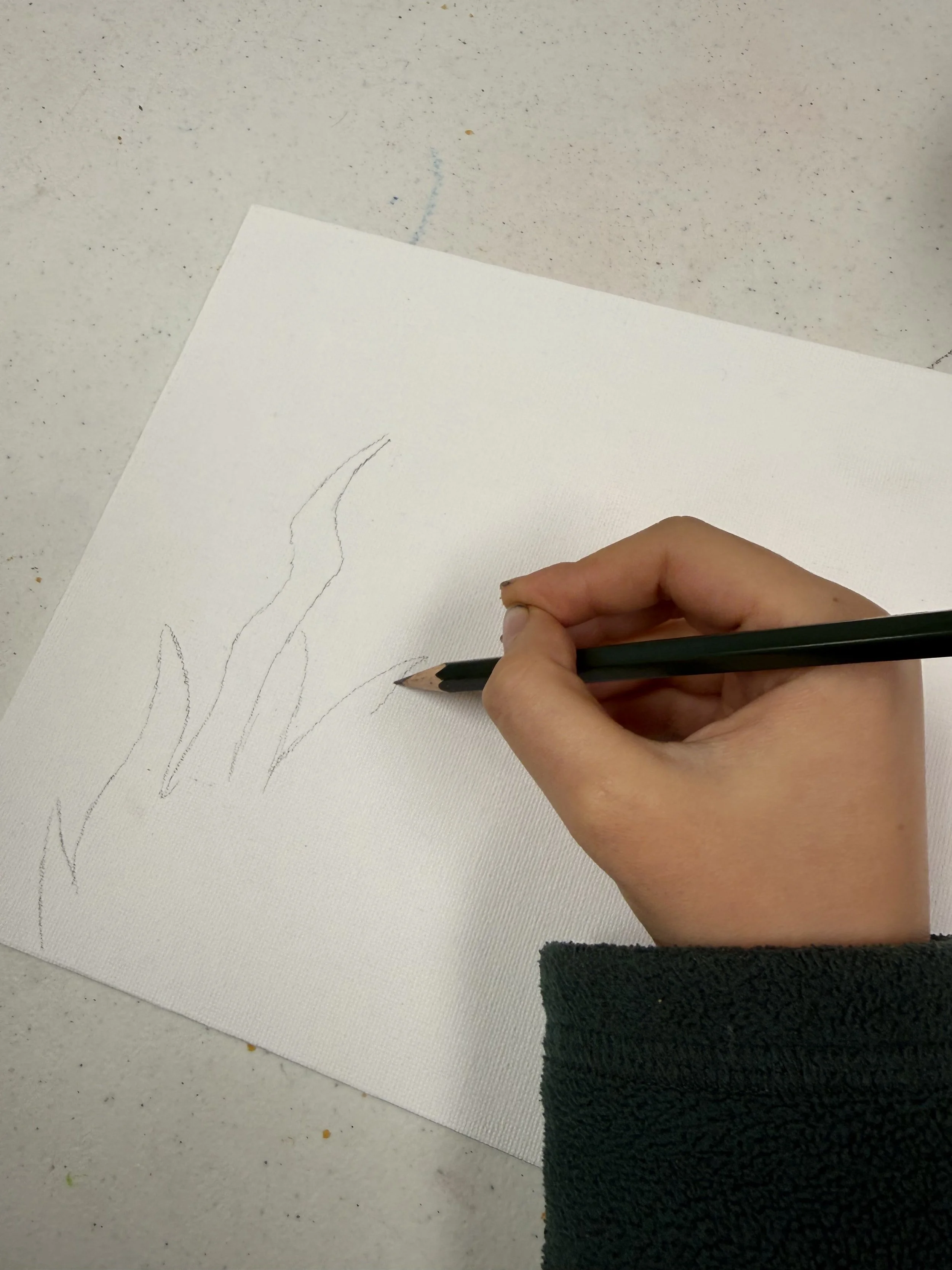 A person drawing on white paper with a black pencil, with a few light pencil lines visible, on a speckled white tabletop.