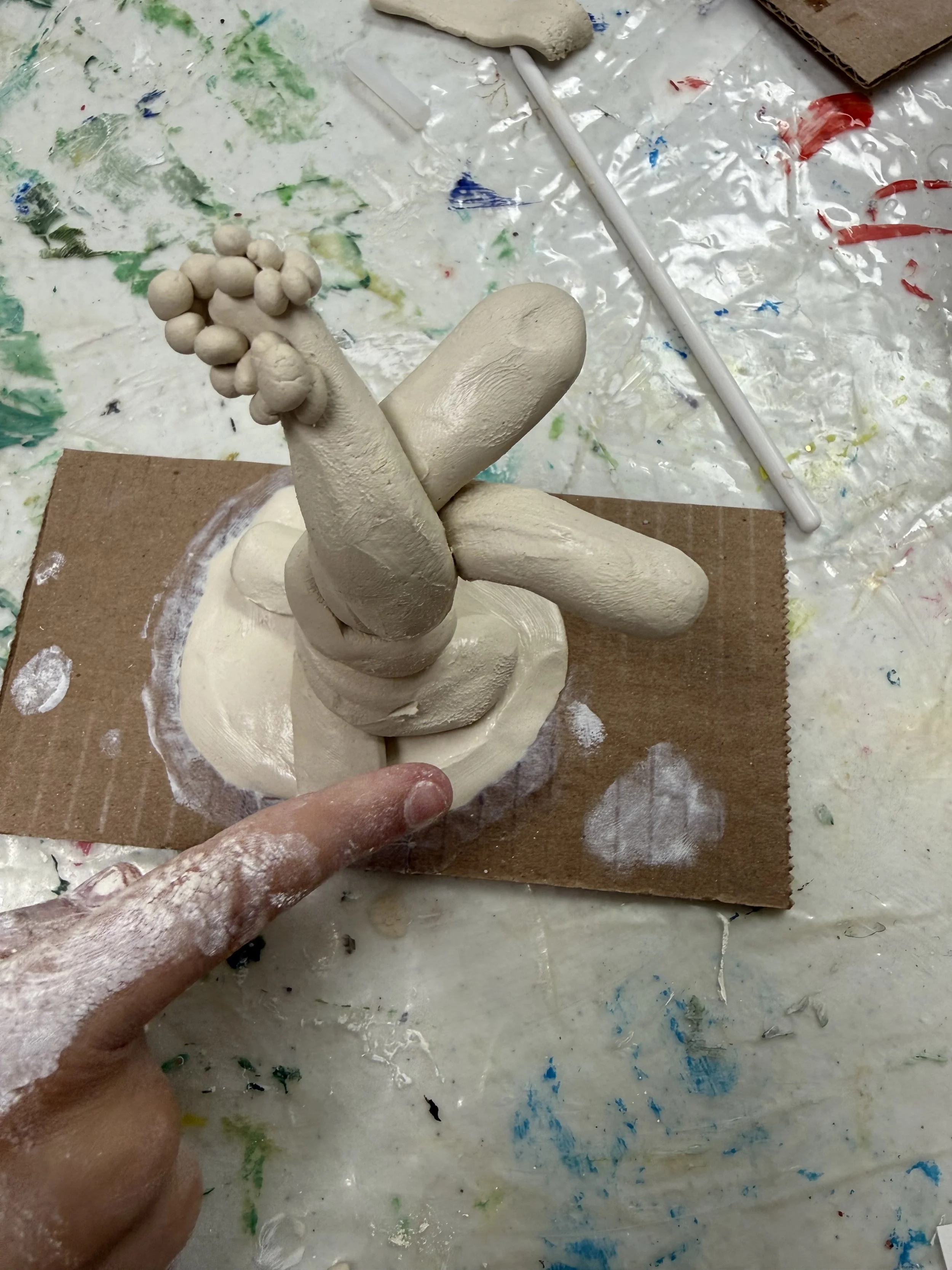Finger pointing to a clay sculpture resembling a cactus with bulbous branches, on a messy art table with paint splatters and tools.