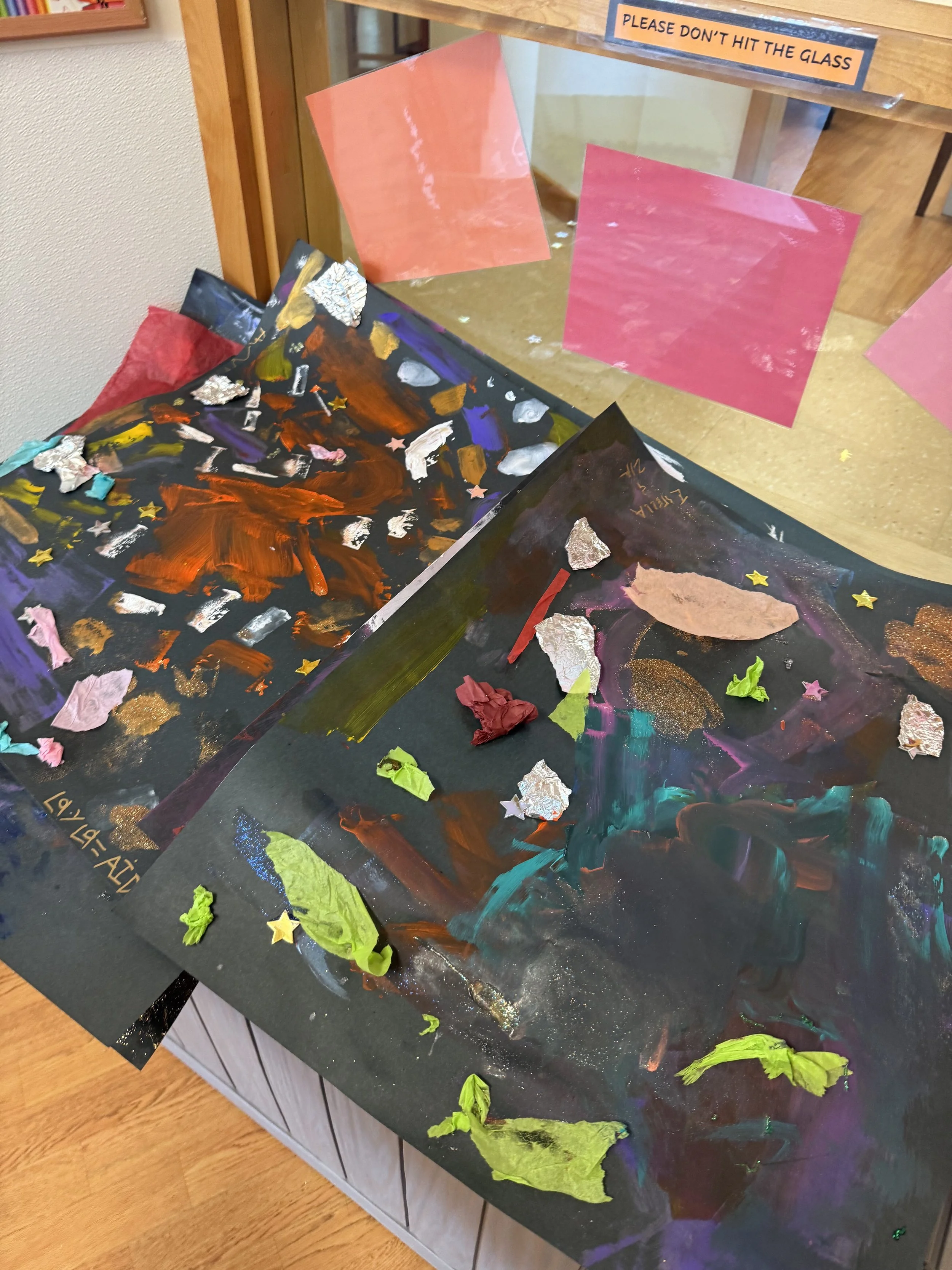 Colorful abstract artwork on black paper with crumpled tissue paper, star-shaped stickers, and paint, displayed on a table near a glass barrier with pink paper and a sign reading 'Please don't hit the glass.'
