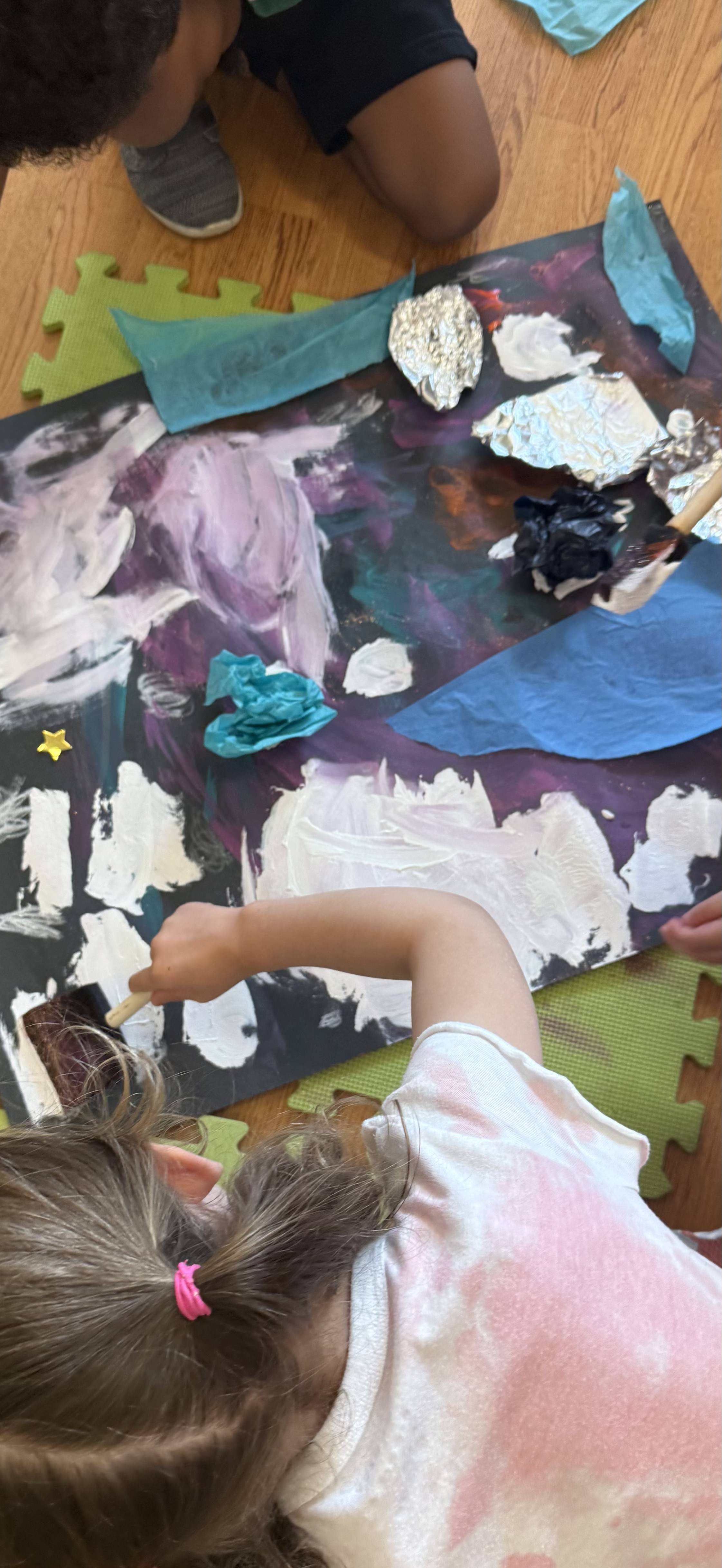 Children working on a space-themed art project with paint, crumpled aluminum foil, tissue paper, and small decorative stars on a black poster board.