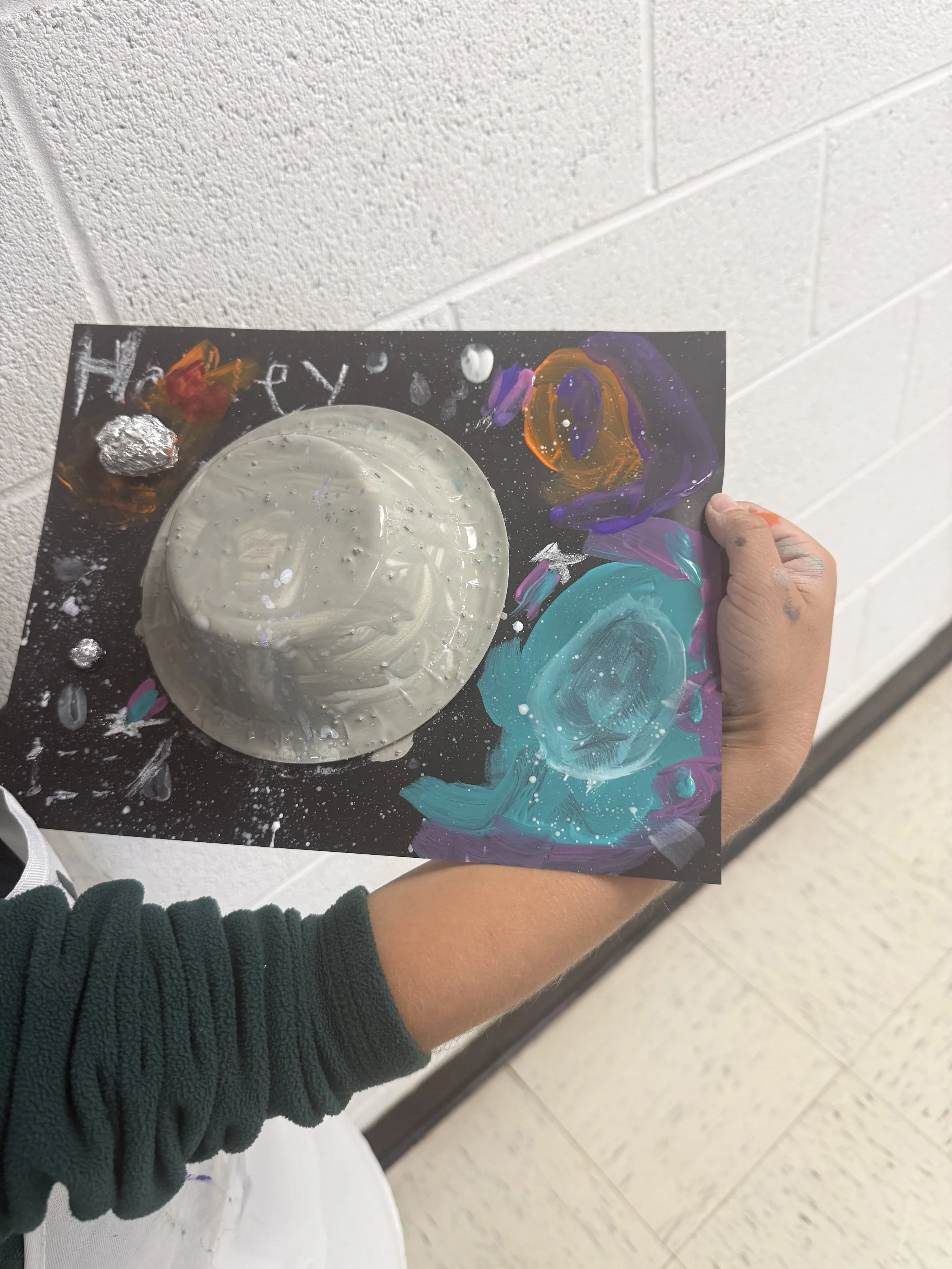 A person holding a painted black sheet of paper with a large grayish-white circular object in the center, surrounded by colorful abstract paint strokes and splatters.