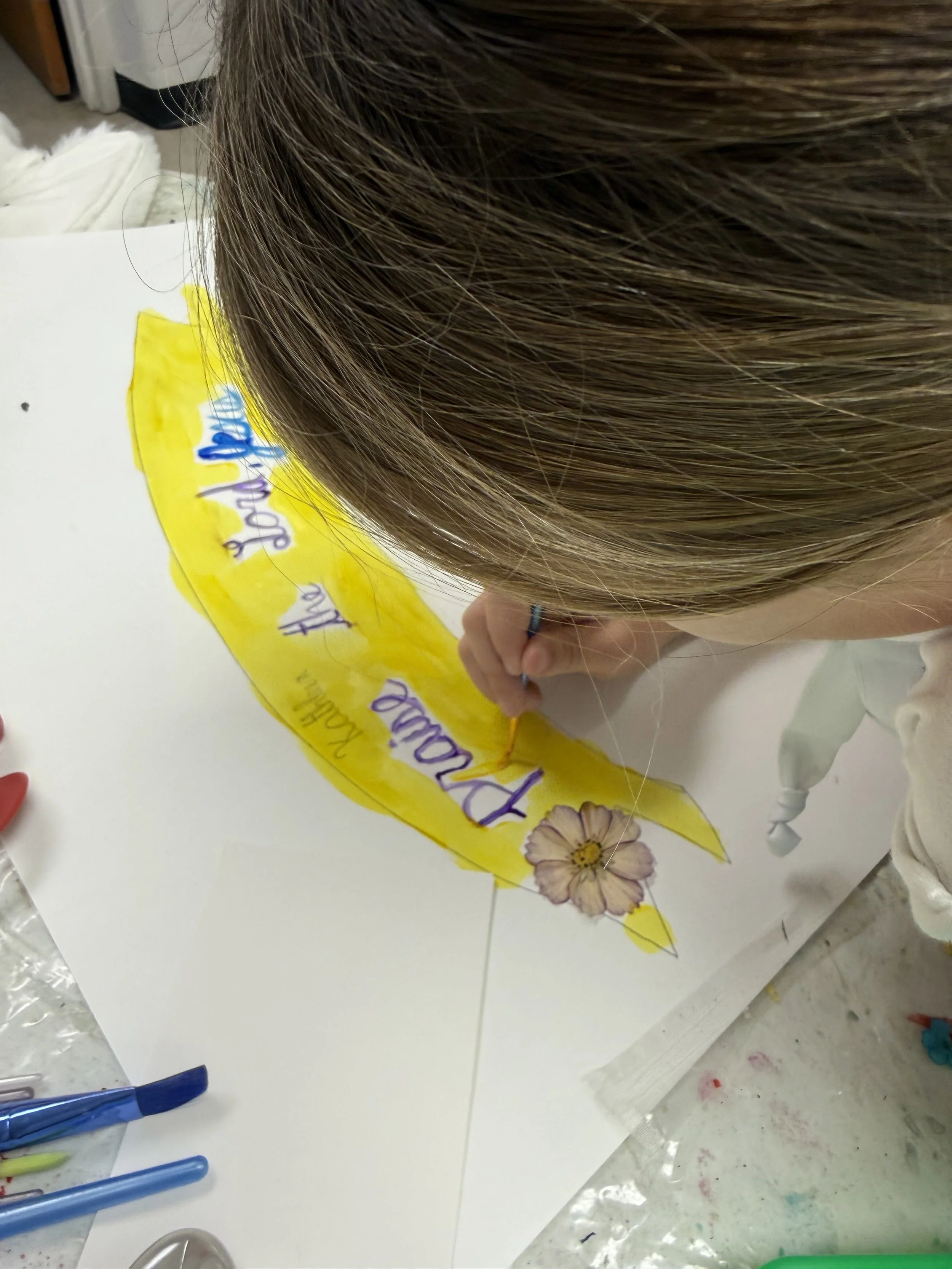 Person coloring a yellow leaf with a flower on it and handwriting on a white sheet of paper.
