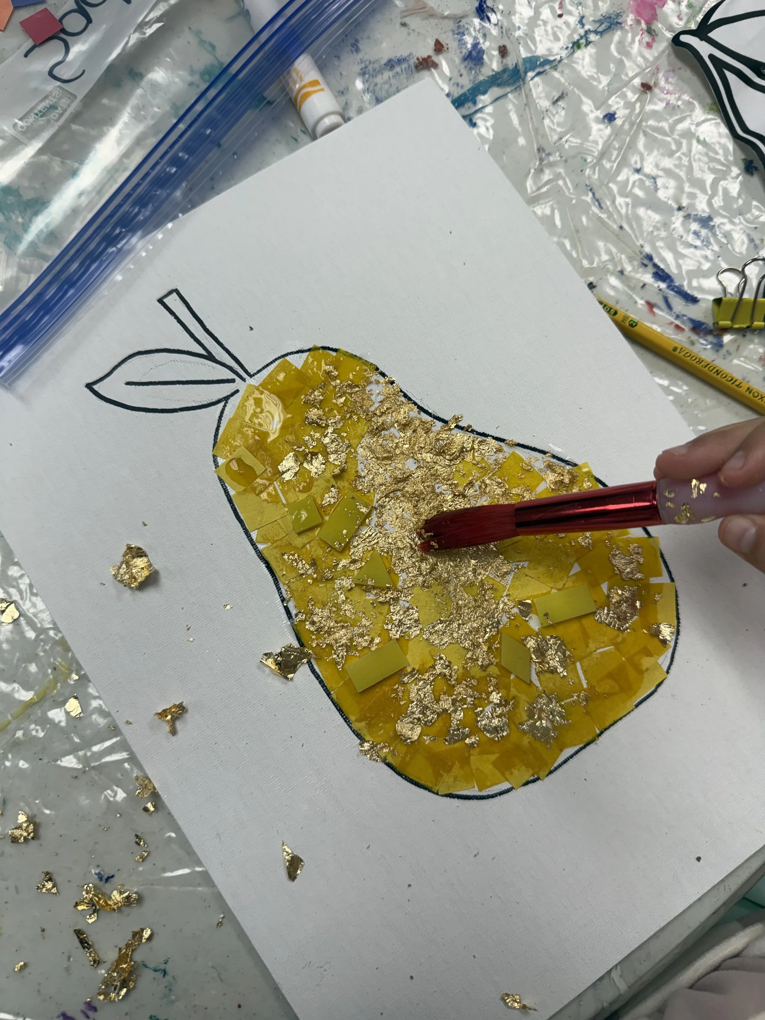 A craft project of a pear shape outline on a canvas with yellow and gold foil pieces being glued onto it, and a hand holding a red glue applicator.