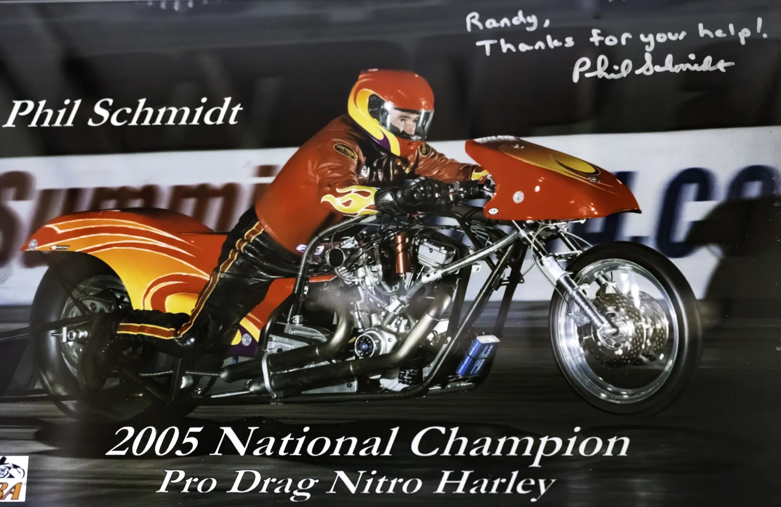 A man in a red racing suit and helmet riding a custom-built red and yellow drag racing motorcycle on a race track. The motorcycle is a Harley Davidson Pro Drag Nitro Harley, and the text indicates he was the 2005 national champion. There is handwritten text thanking Randy for help and signed by Phil Schmidt.