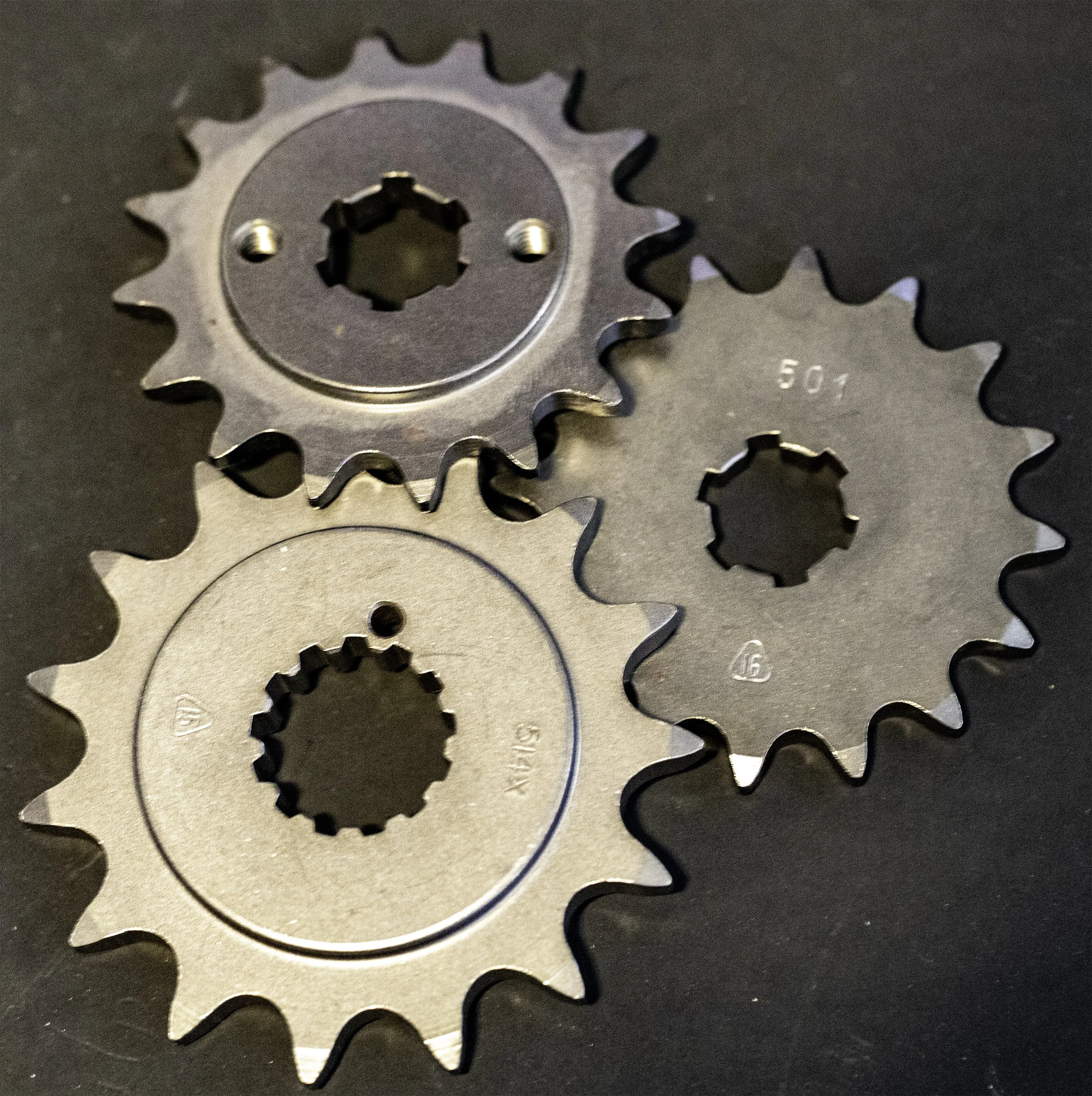 Three metal motorcycle sprockets with teeth, placed on a dark surface.