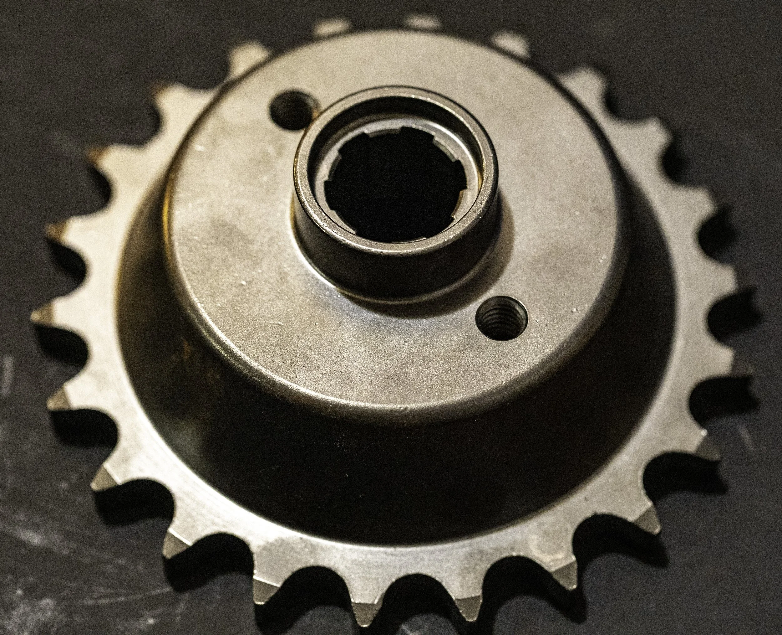 A metal motorcycle sprocket with teeth, the center hole, and two smaller screw holes, resting on a dark surface. Harley Davidson