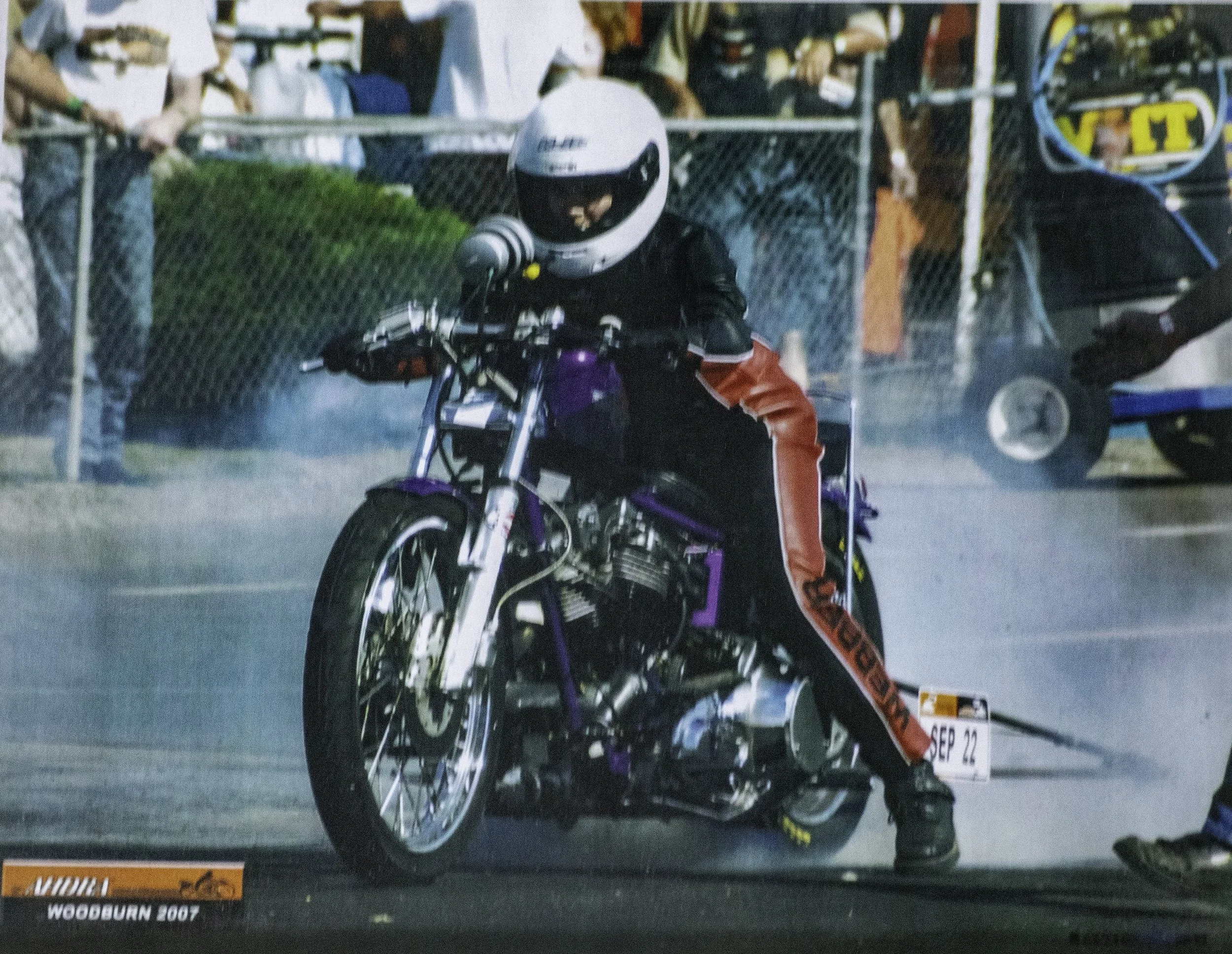 A person wearing a white helmet, black jacket, and red racing pants performing a burnout on a purple motorcycle at a drag race event. Smoke is coming from the rear tire, and spectators are visible behind a fence in the background.