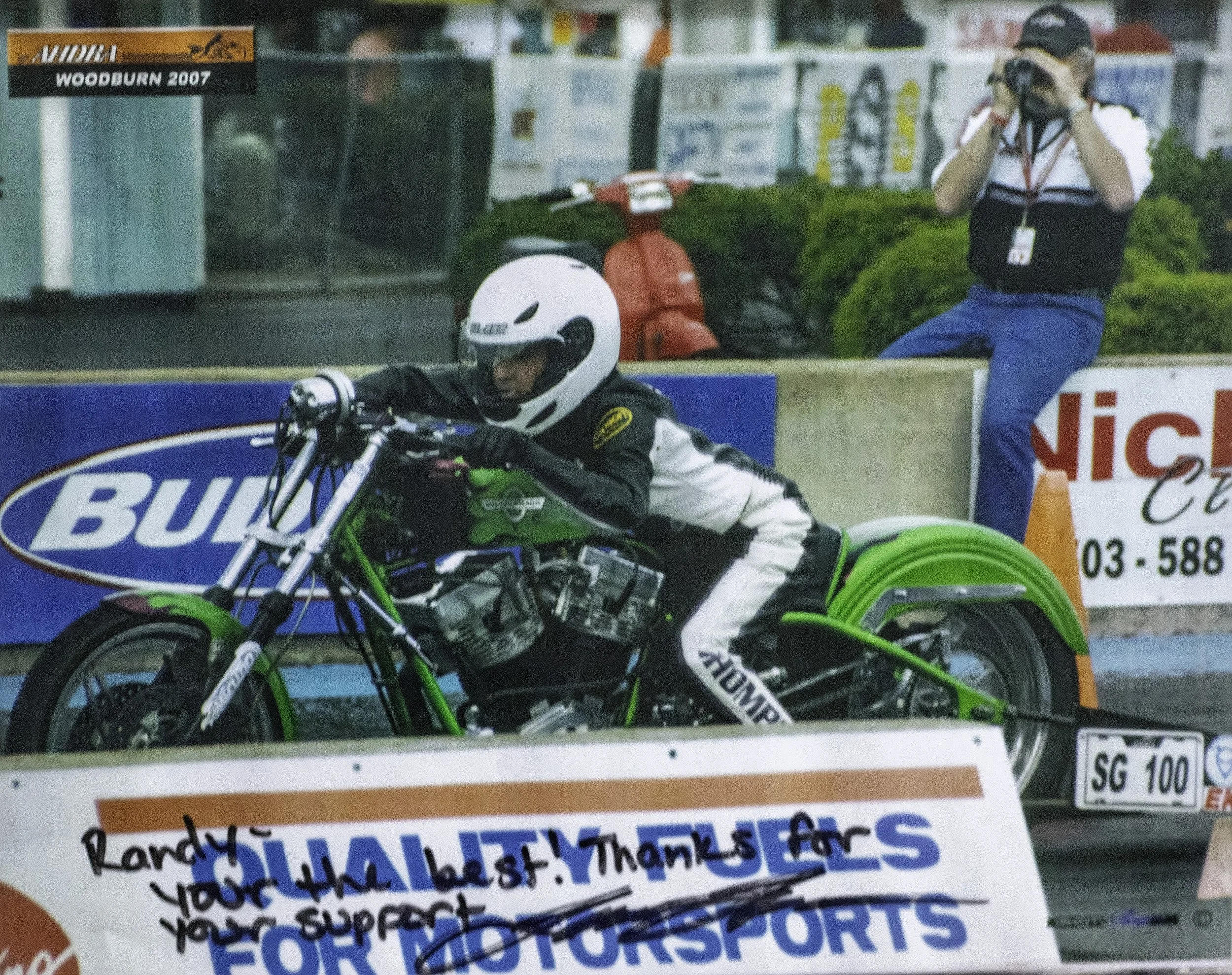 A person dressed in racing gear riding a green drag bike during a race event. A photographer is taking pictures in the background, and there are banners and tents nearby.