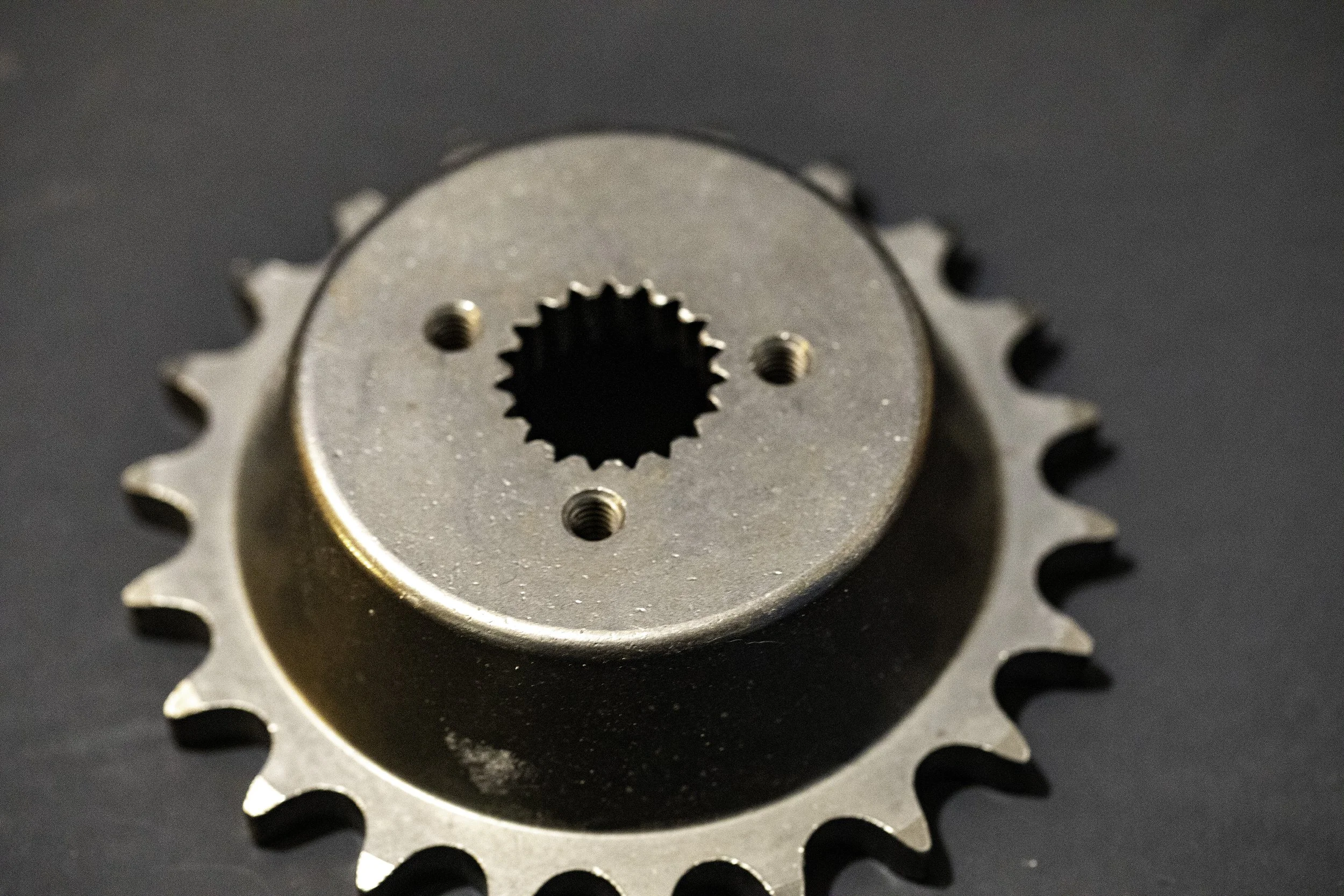 A close-up of a metal bicycle sprocket or gear with teeth around its edge, sitting on a dark surface.