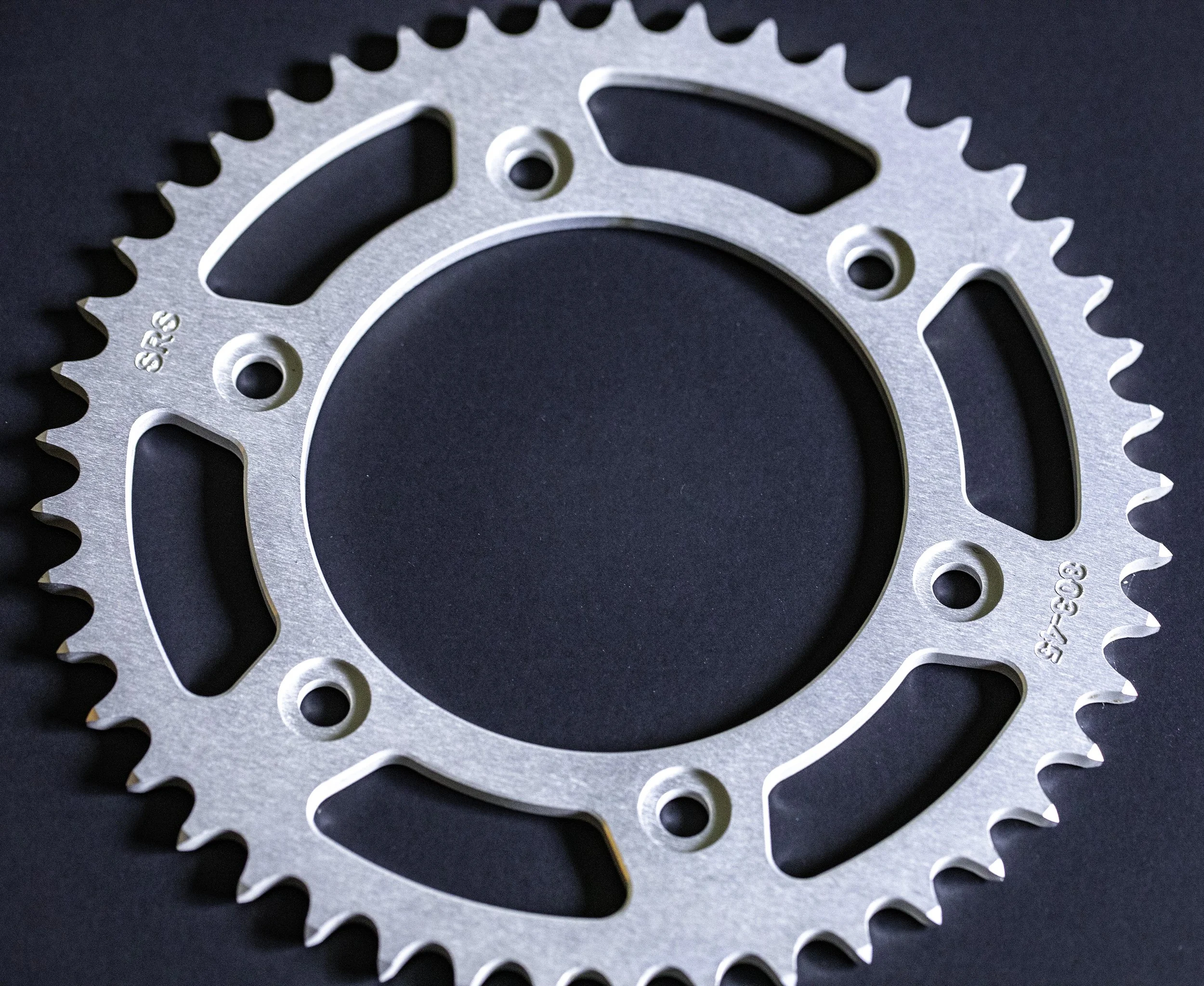 Close-up of a silver motorcycle rear sprocket with multiple teeth on a black background.