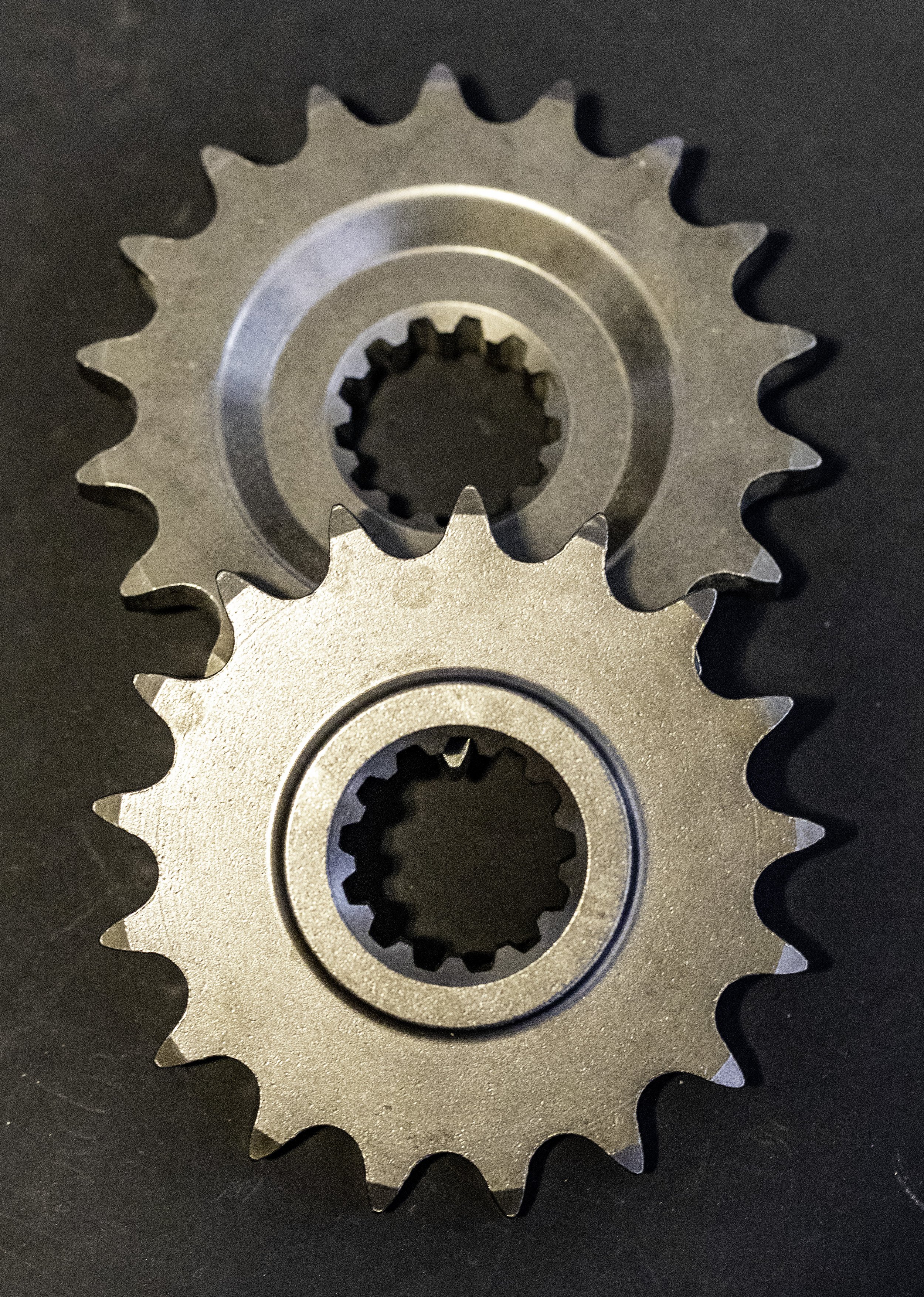 Two metal sprockets with central splined holes, placed on a dark surface.