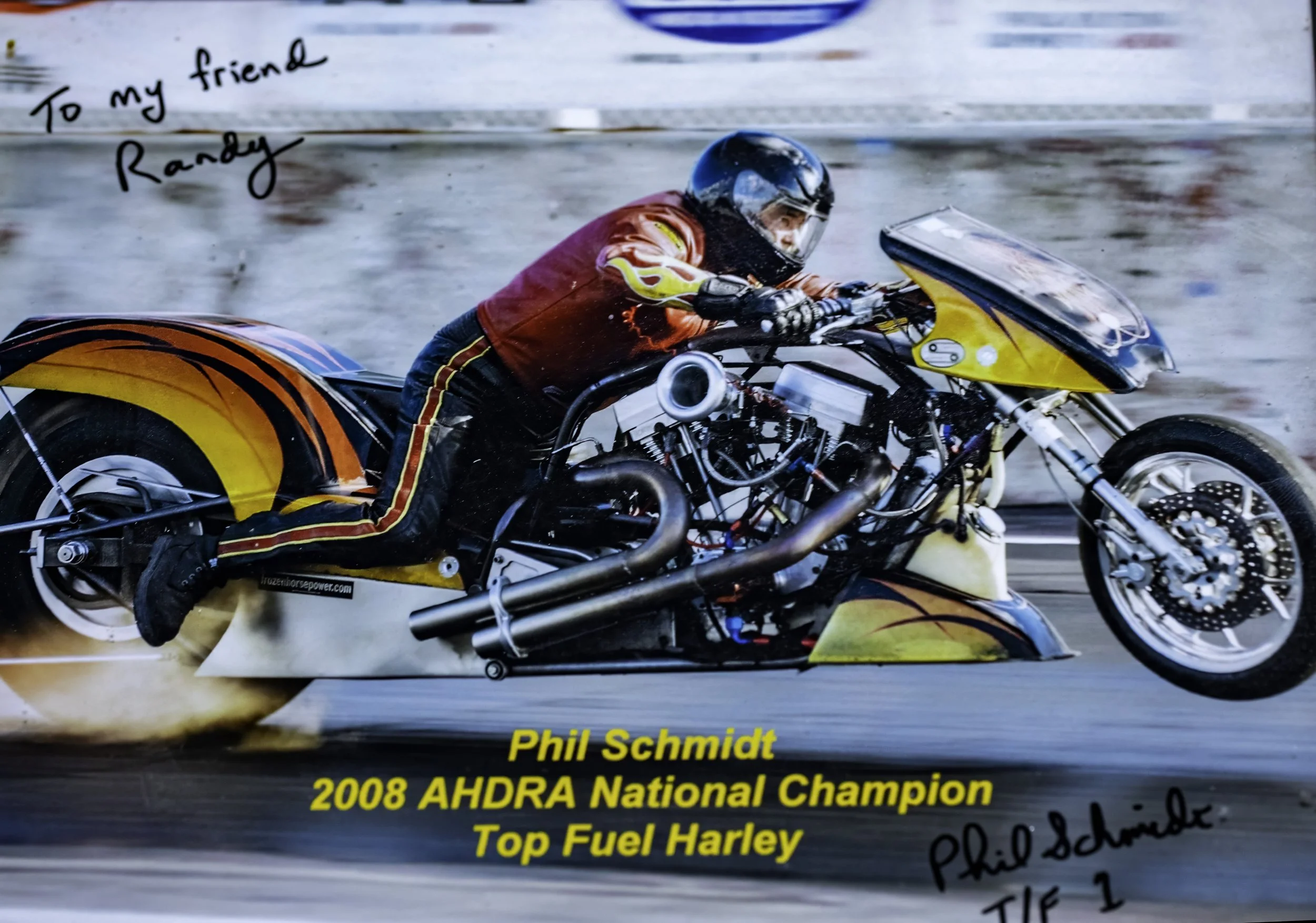 A person riding a top fuel Harley-Davidson motorcycle at high speed, wearing a helmet and red racing suit, with a handwritten note and signature in the corner.