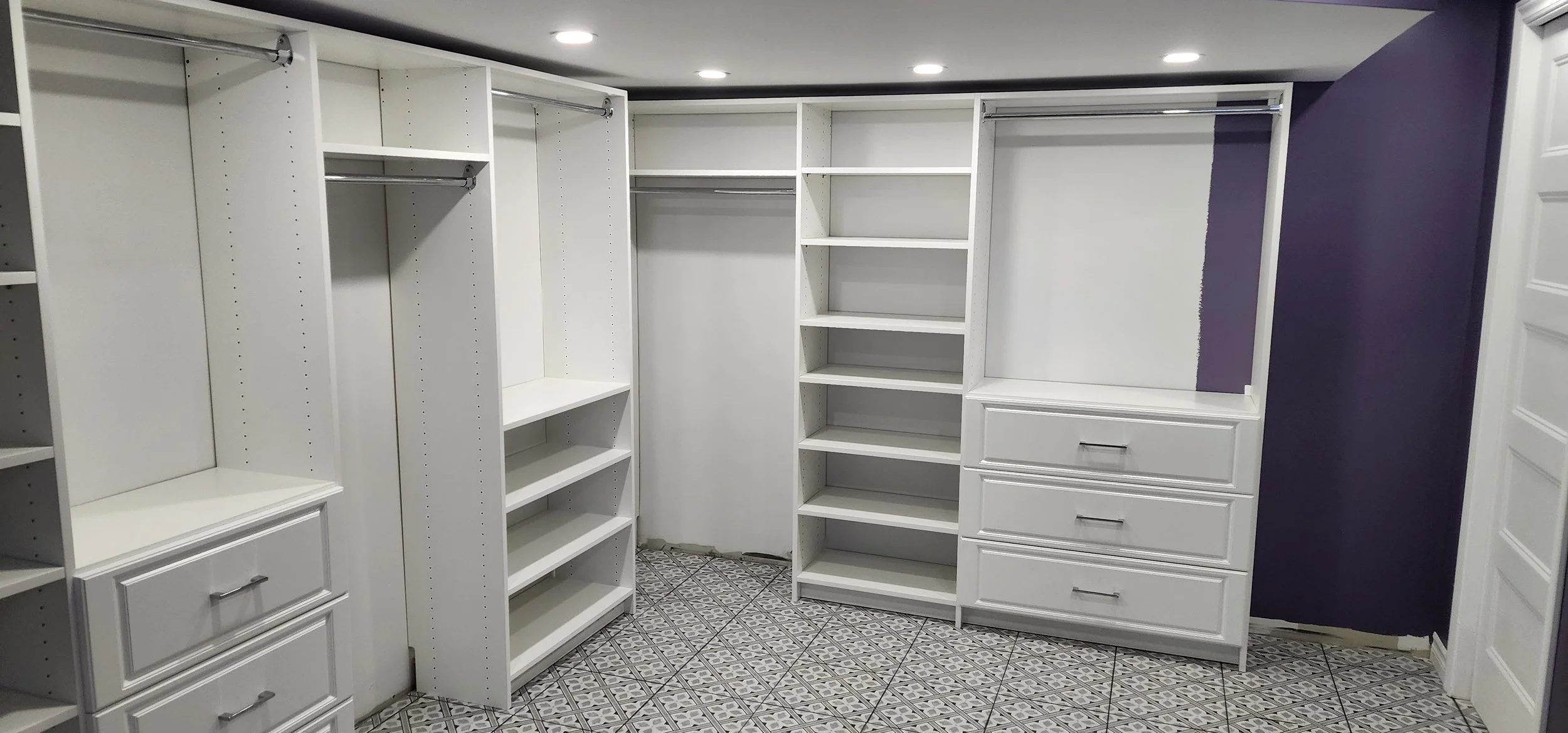 Empty walk-in closet with white shelving units, drawers, and hanging rods, purple accent wall, patterned tile floor, and ceiling lights