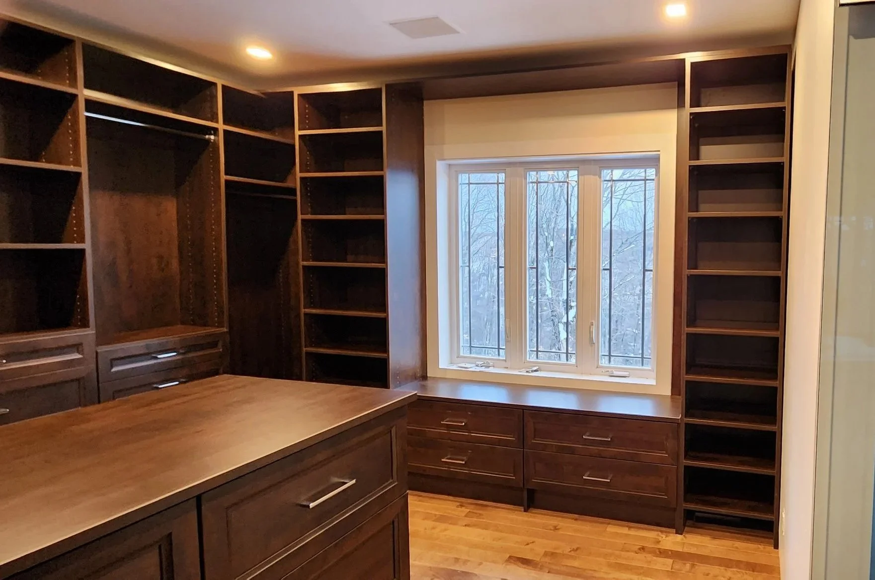 Empty custom-built dark wood shelving and cabinets in a room with hardwood floors and a large window.