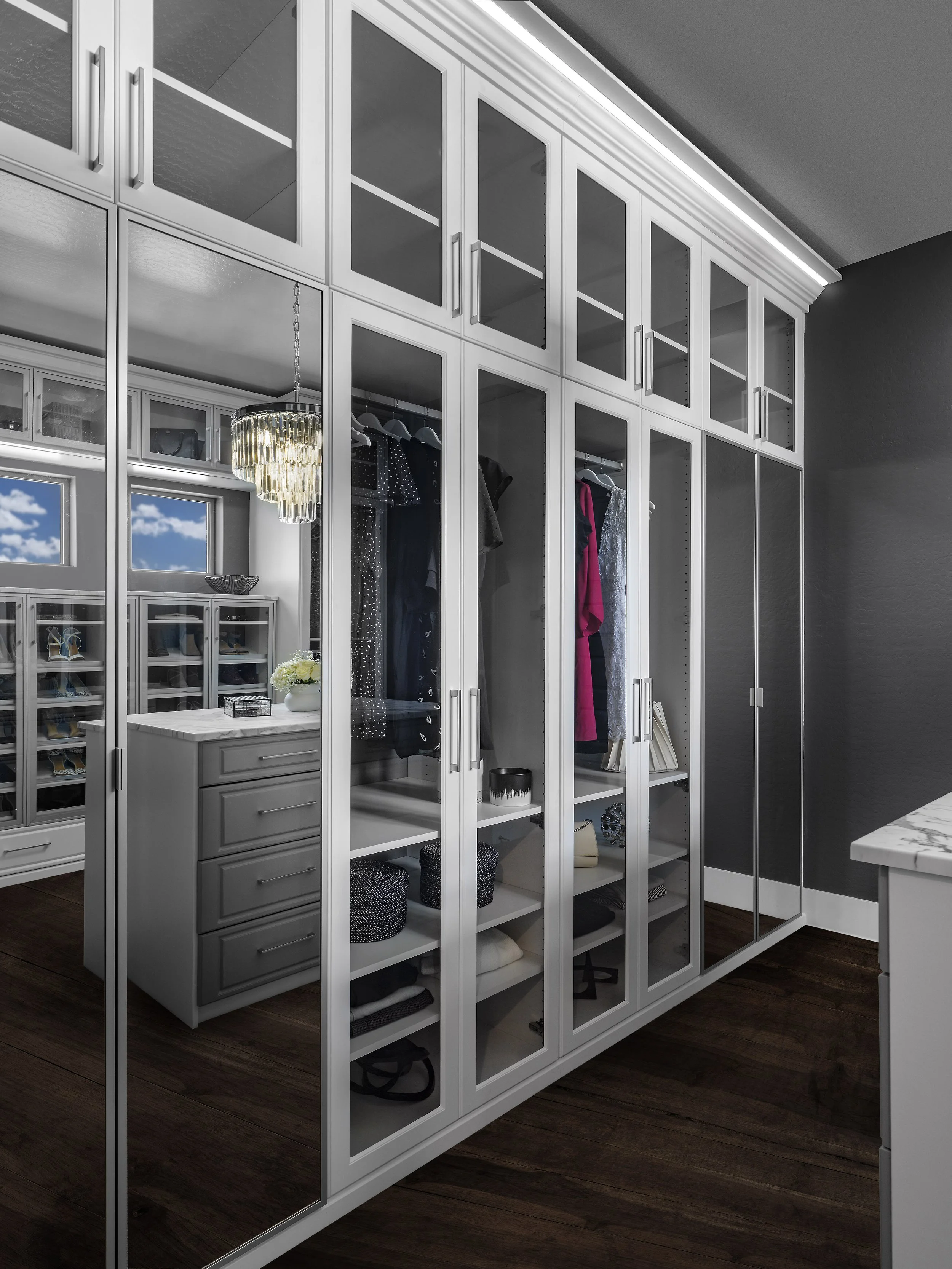 Modern walk-in closet with white cabinets, glass doors, and built-in shelves, containing hanging clothes, storage baskets, and a jewelry display, with dark wood flooring.