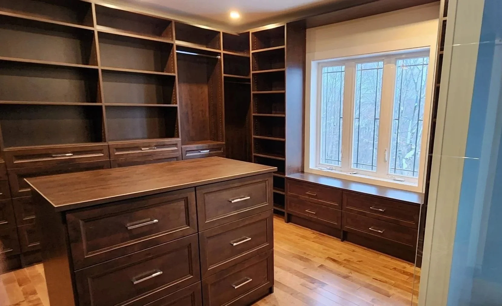 A room with custom dark wood shelving and cabinetry, a central island with drawers, and a large window with a wooden bench below, overlooking a wooded outdoor scene.