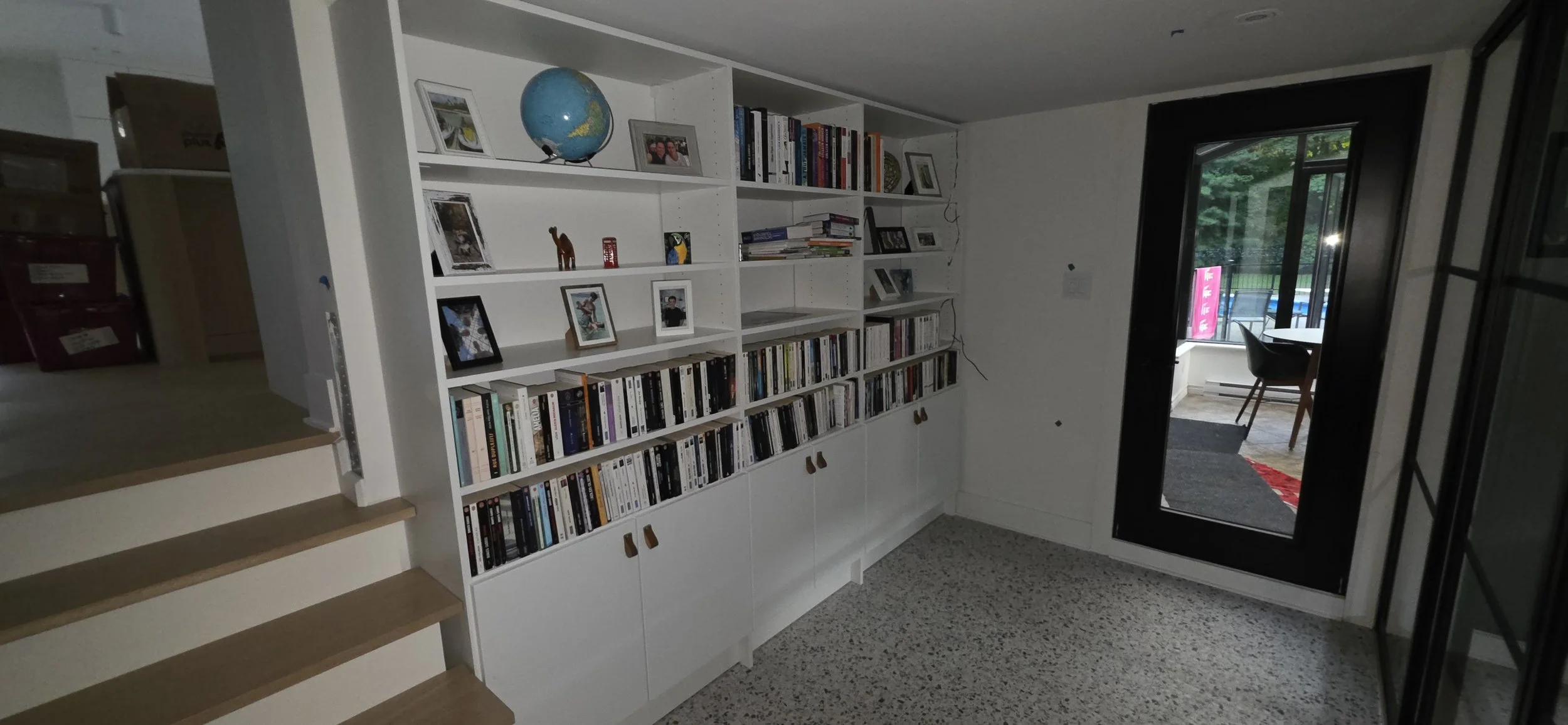 Interior view of a modern living space with a white bookshelf filled with books, framed photos, and decorative items. There is a doorway with black framing leading to a sunlit outdoor patio area with a table and chairs, and a large window showing greenery outside.
