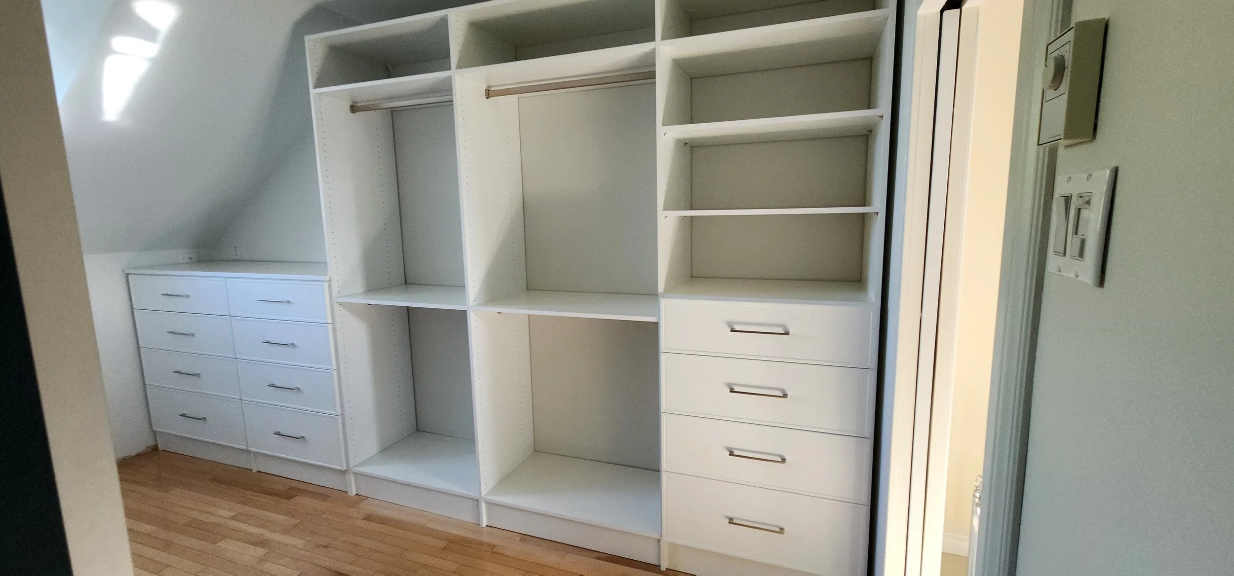 Empty built-in white closet with five sections, some with shelves and some with hanging rods, on a wooden floor with a sloped ceiling. There are electrical switches and outlets on the right wall.