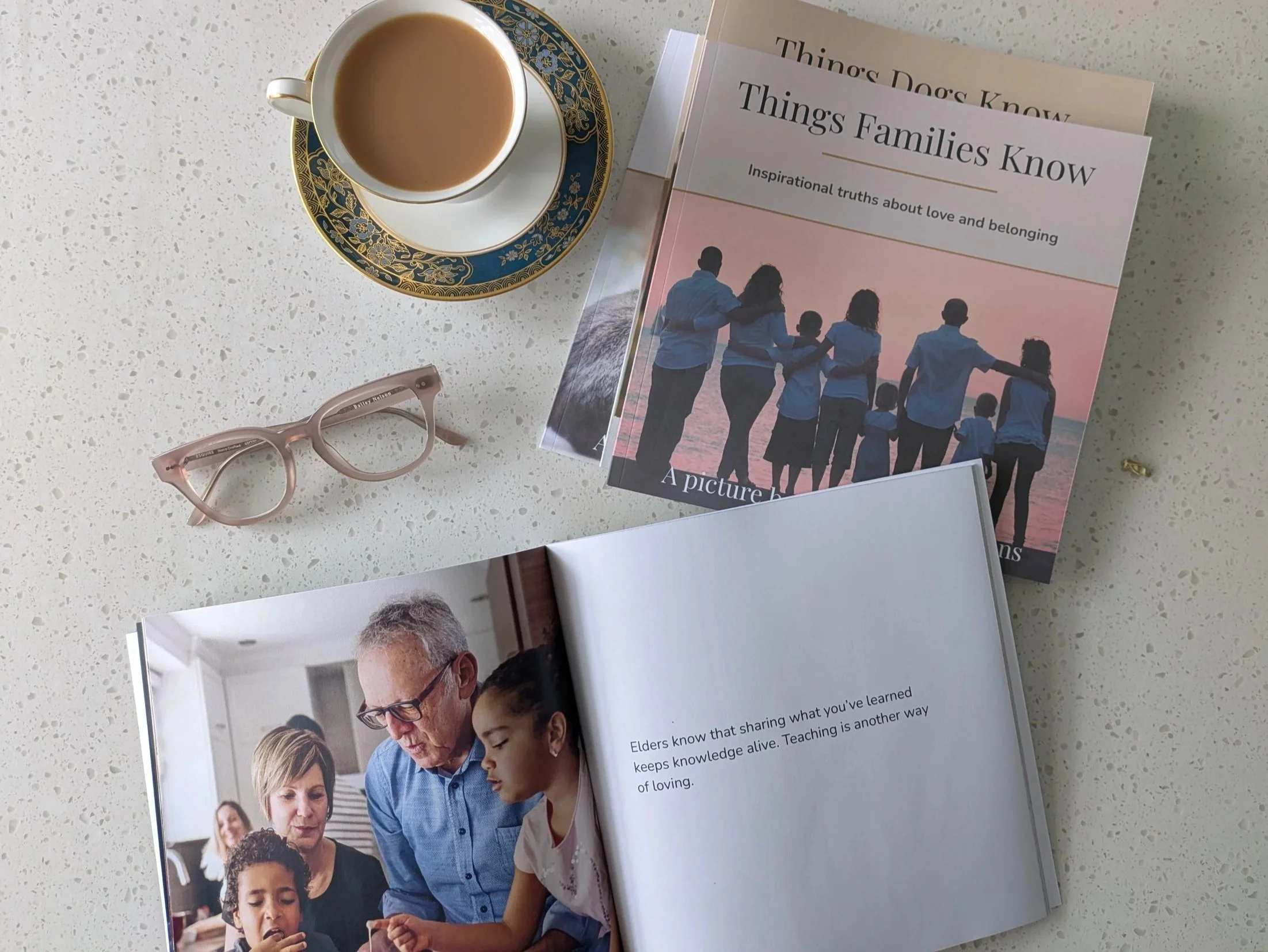 A collection of teal-colored books titled "Things Dogs Know" and "Things Families Know", a cup of coffee on a decorated saucer, a pair of pink eyeglasses, and an open book with a photo of a diverse group of people including children and elders sharing a moment together.