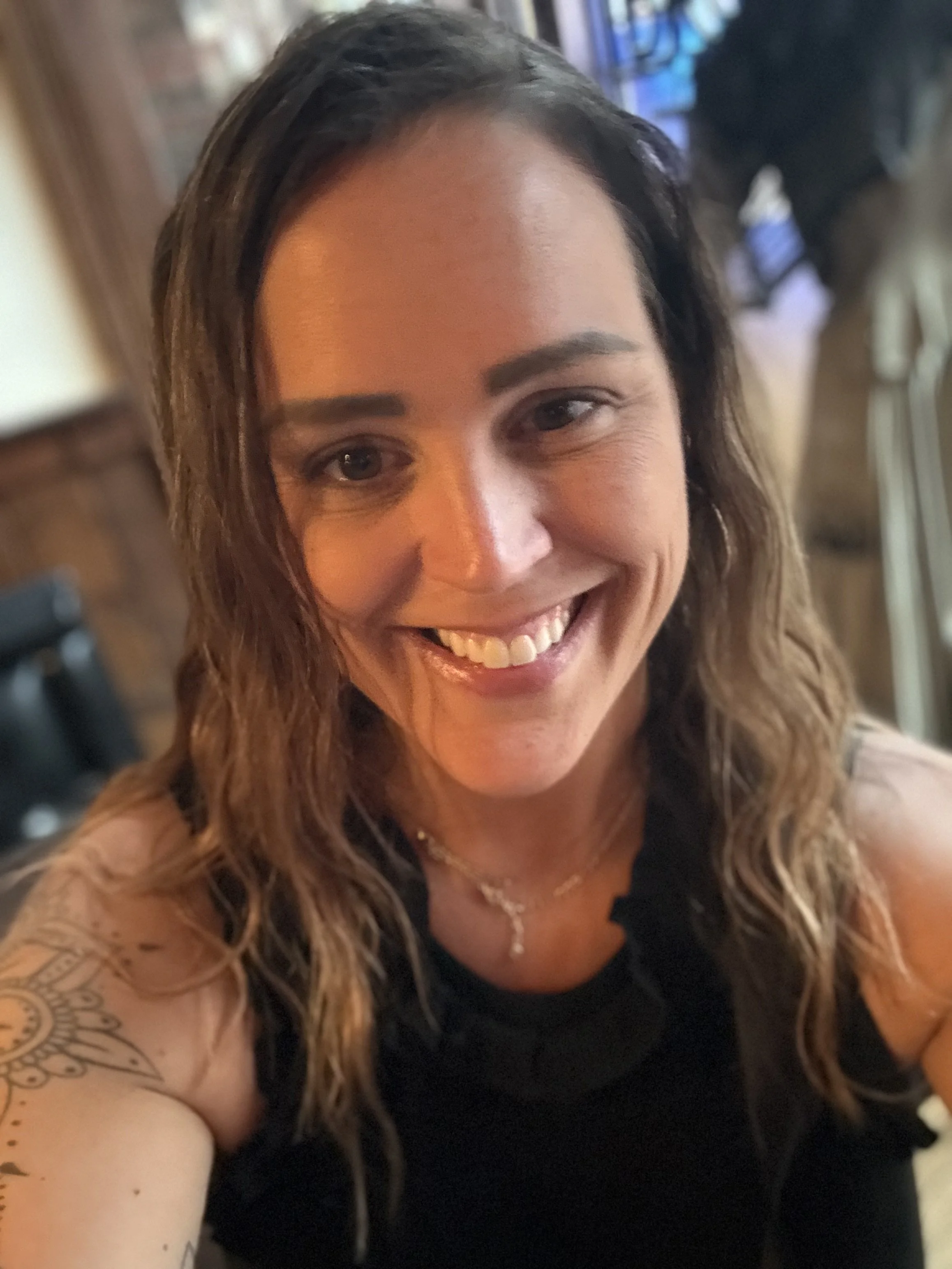 A woman with wavy brown hair smiling for a selfie, showing her teeth, wearing a black top and a necklace, with a tattoo on her upper arm, in an indoor setting with warm lighting.