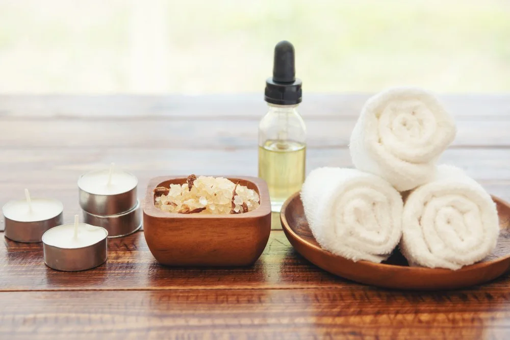 Massage setting with white rolled towels, candles, a small dropper bottle, and bath salts on a wooden surface.