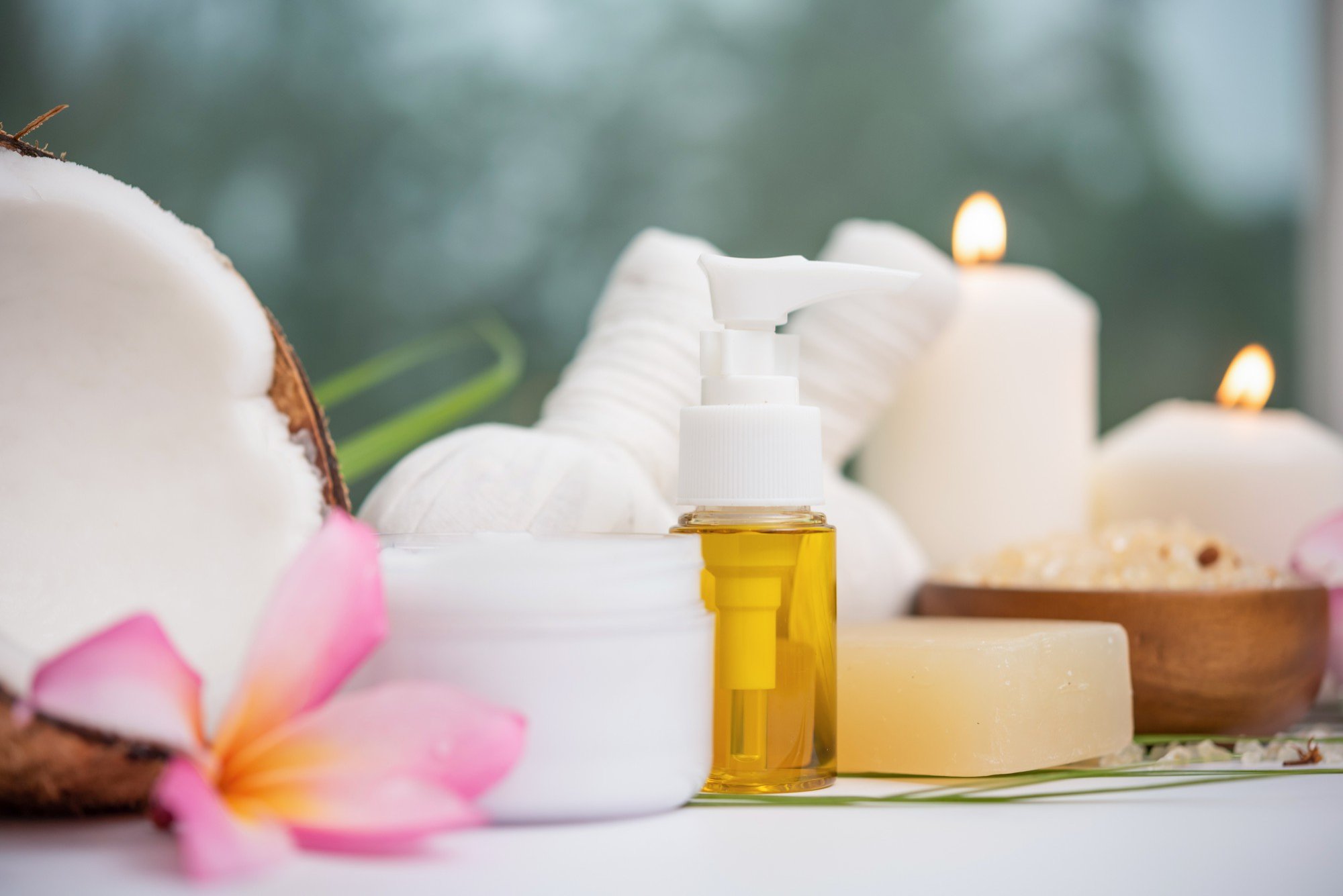 Wellness setup with coconut shells, pink flowers, candles, a small bottle of massage oil, and soap on a table.