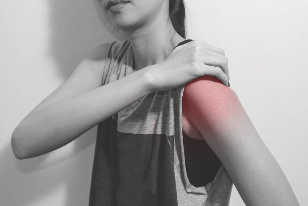 A woman in a sleeveless shirt, touching her inflamed and painful shoulder, indicating pain or injury.