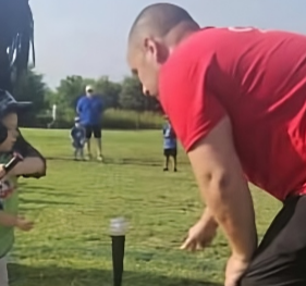 A man in a red shirt bends over a table with a cup on it, while a young girl watches, outdoors on a grassy field with several people and trees in the background.