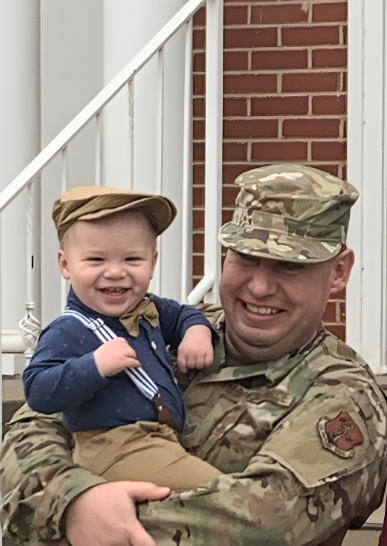 A soldier in uniform holding a smiling young boy dressed in vintage-style clothing, on a porch with a brick wall and white railing.
