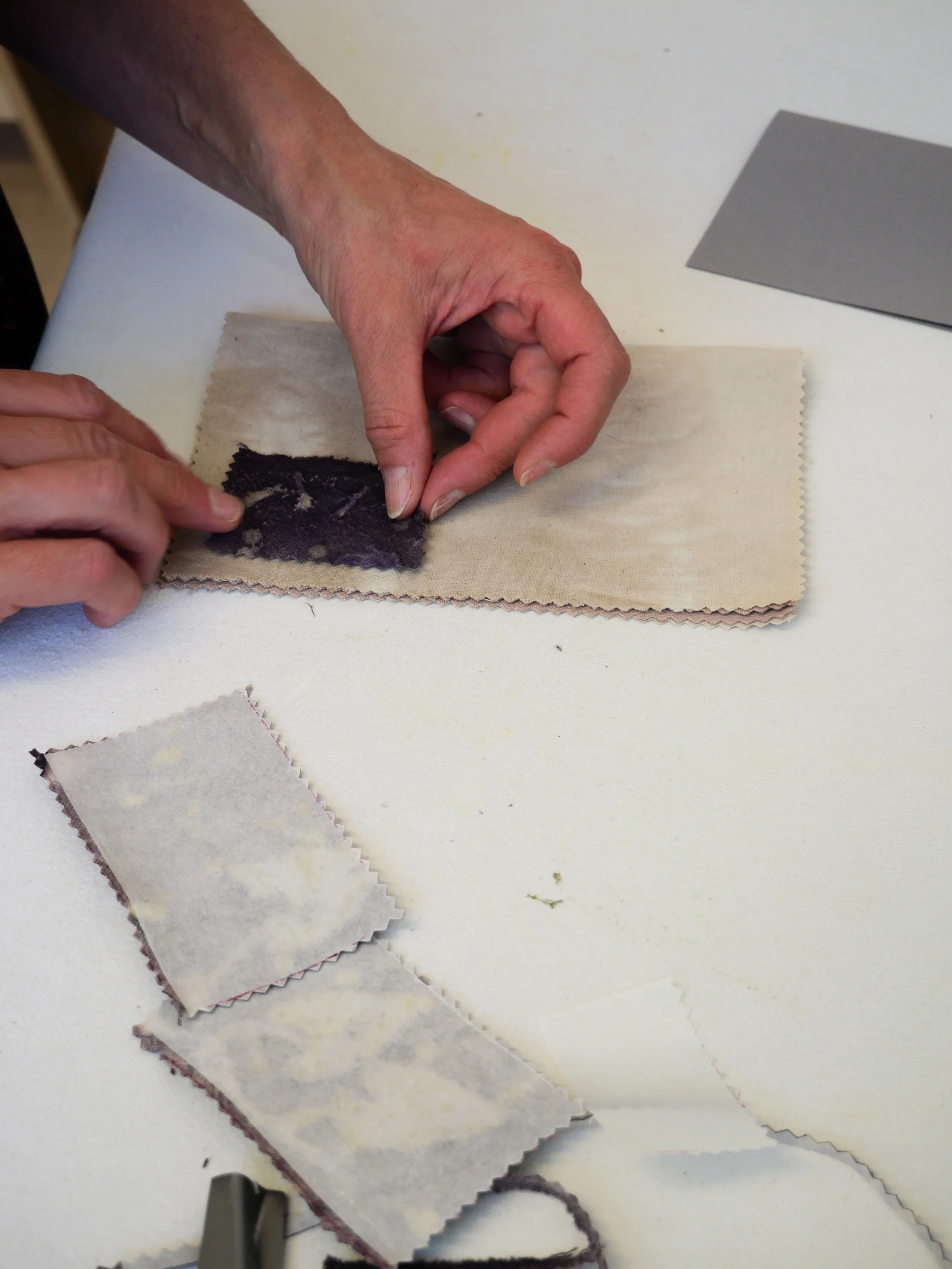Une personne manipule un échantillon de tissu ou de matériau textile, avec d'autres morceaux de tissu autour, sur une table blanche.