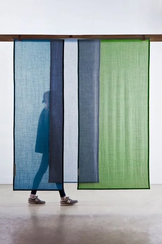 Une personne se tient derrière un mur de rideaux en tissu colorés, avec des couleurs allant du bleu au vert, dans une galerie d'art.