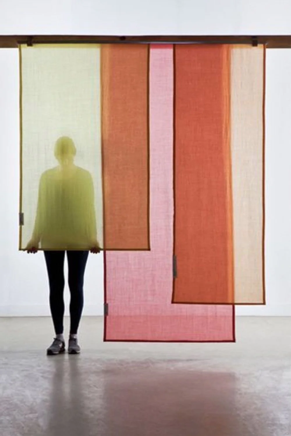 Une personne debout derrière des panneaux translucides de couleur jaune, rose et orange, dans une pièce avec un mur blanc, la personne est de profil avec des vêtements sombres et des chaussures de sport.