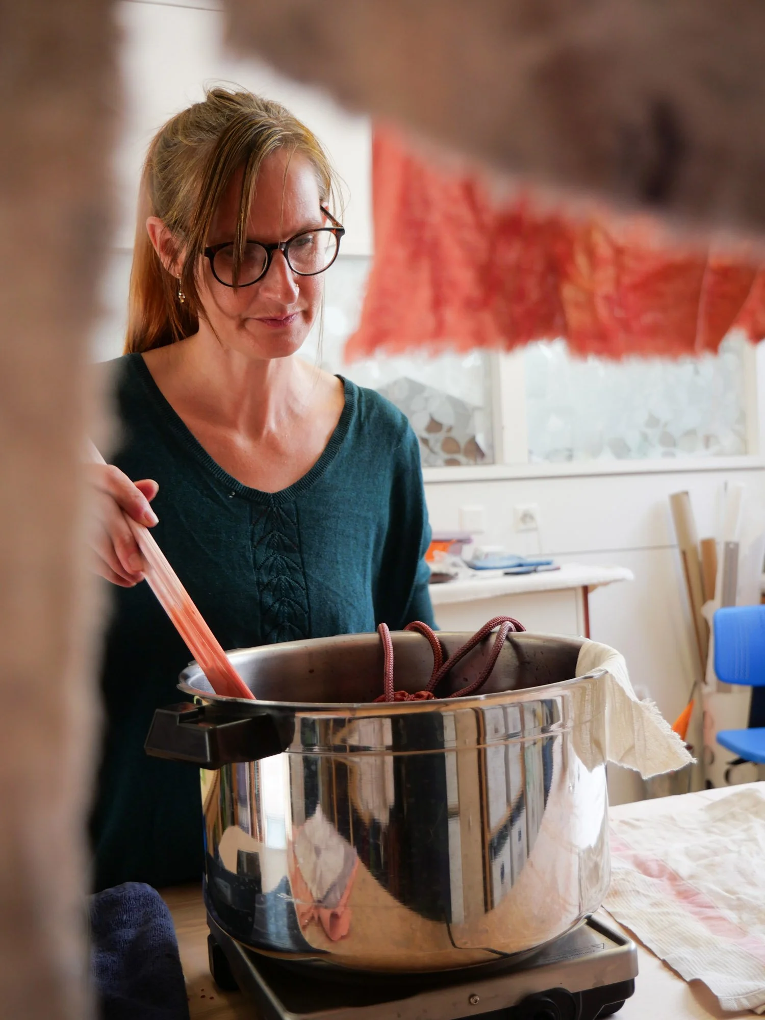 Une femme avec des lunettes, de longs cheveux roux, regarde dans une marmite en métal posée sur une table avec un tissu blanc. Il y a des morceaux de tissu suspendus au-dessus, dans un environnement intérieur lumineux.